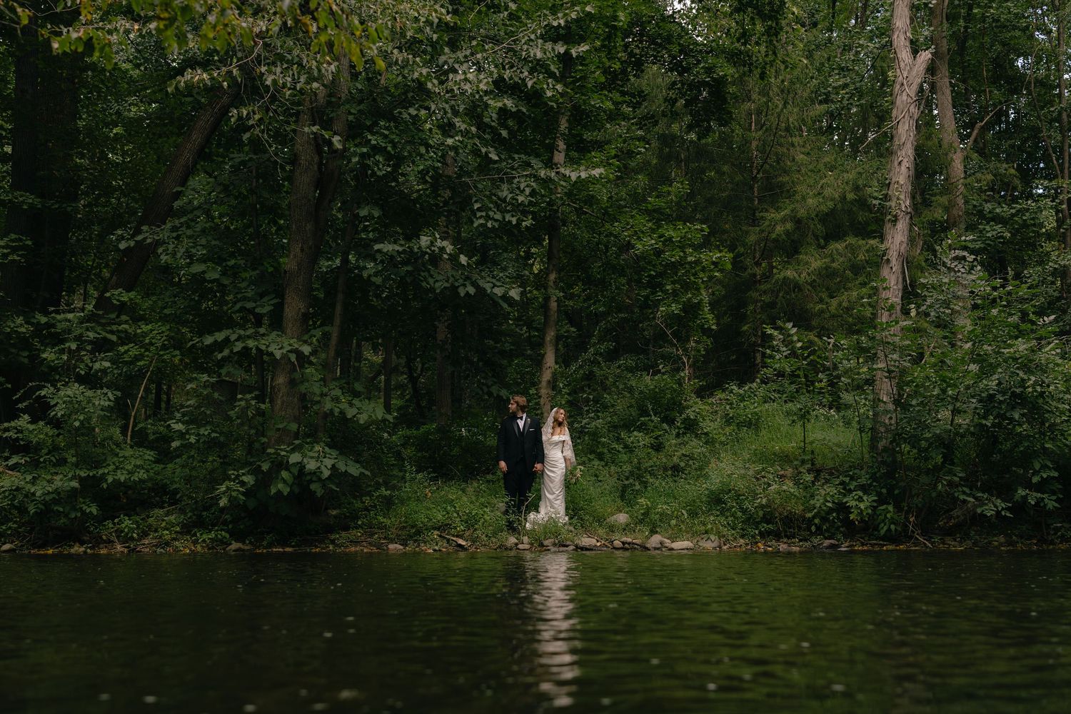 A bride and groom stand together on a serene lakeside forest path surrounded by lush green trees.