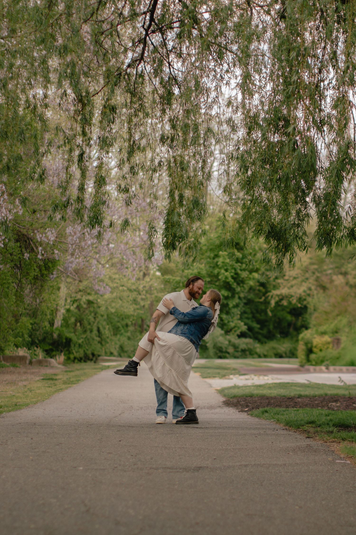 Couple dancing romantically on a park path under a weeping willow tree, surrounded by lush green foliage.