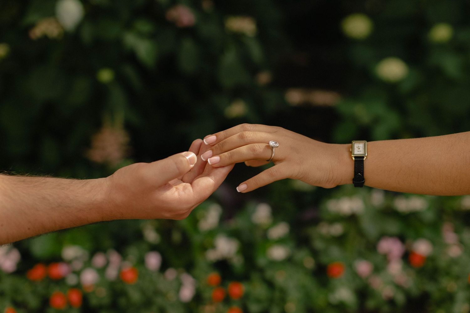 Two hands gently touching outdoors, one wearing a ring and bracelet, with colorful flowers in the background.
