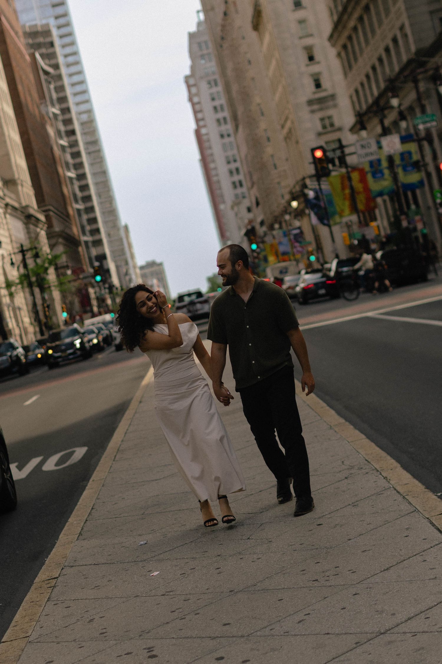 Couple holding hands and walking on a city sidewalk, woman in white dress and man in dark outfit, urban buildings in background.
