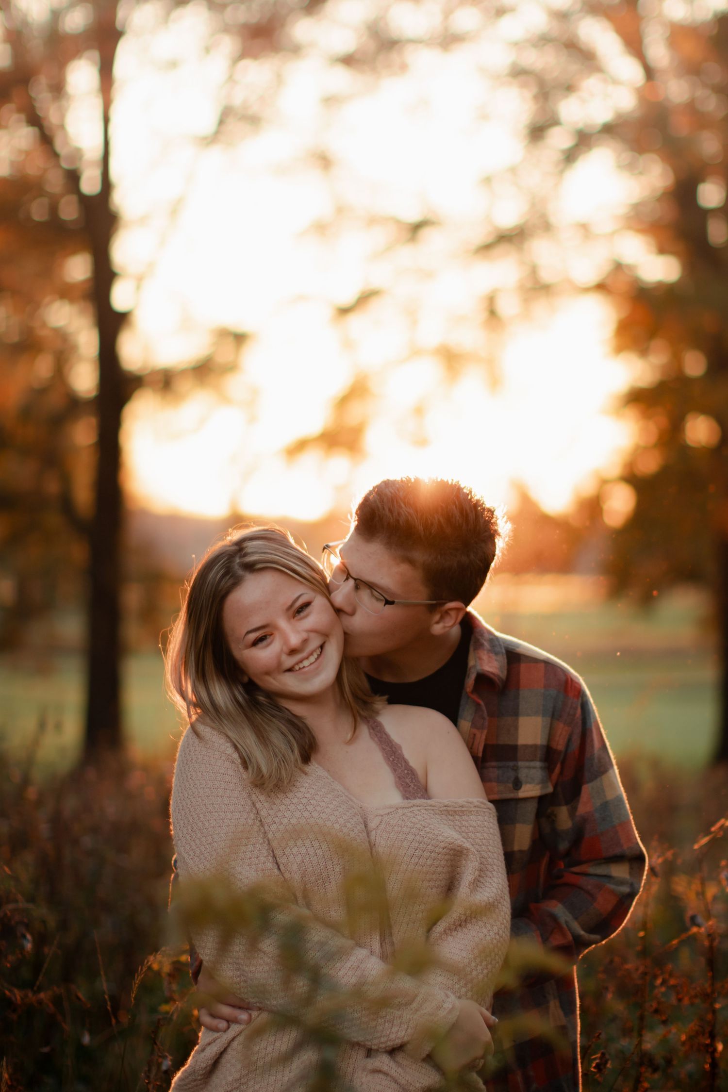 A couple embracing during golden hour in a field with autumn colors and sunlight filtering through trees.