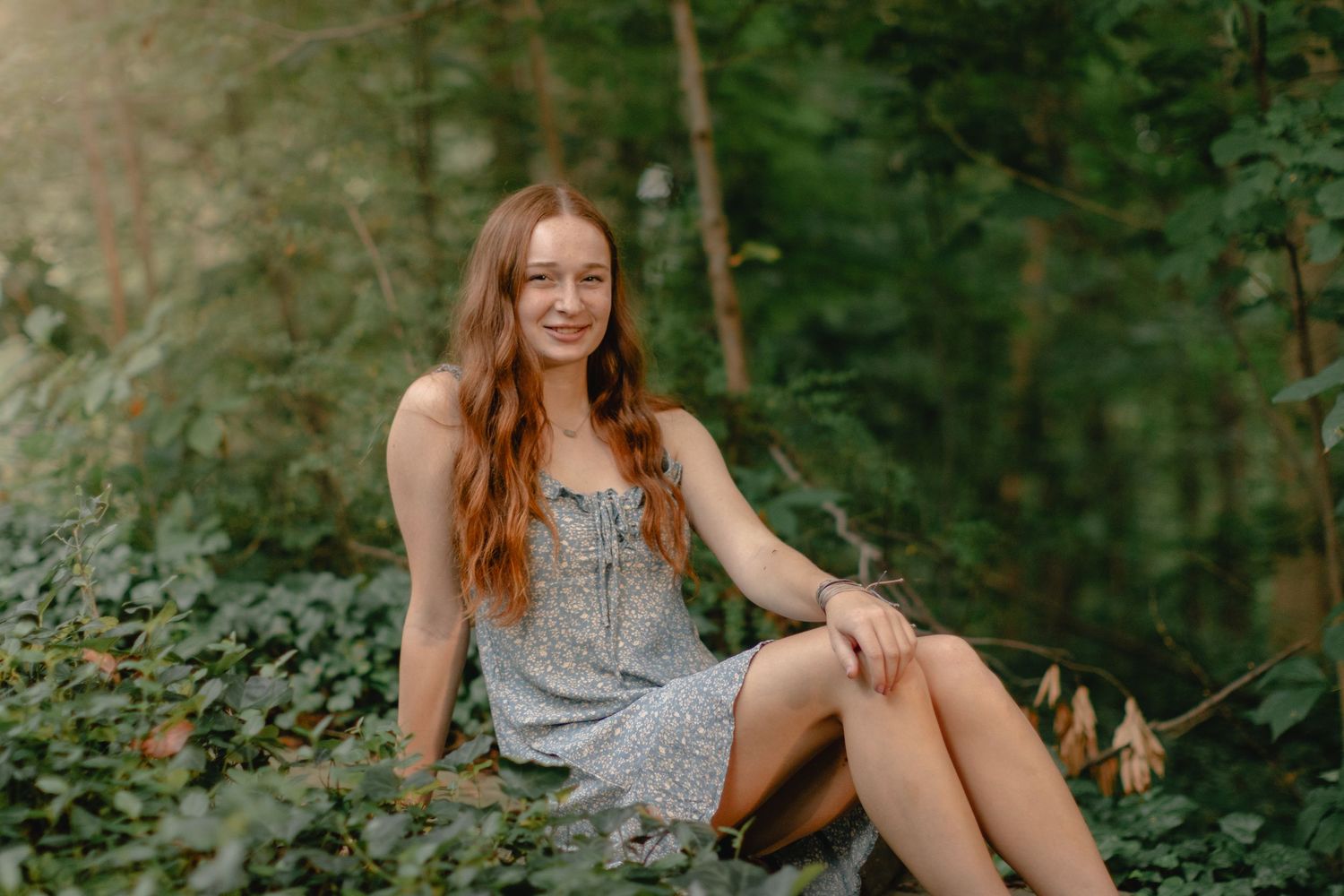 A woman in a floral dress sits on a forest path surrounded by lush green foliage in natural lighting.