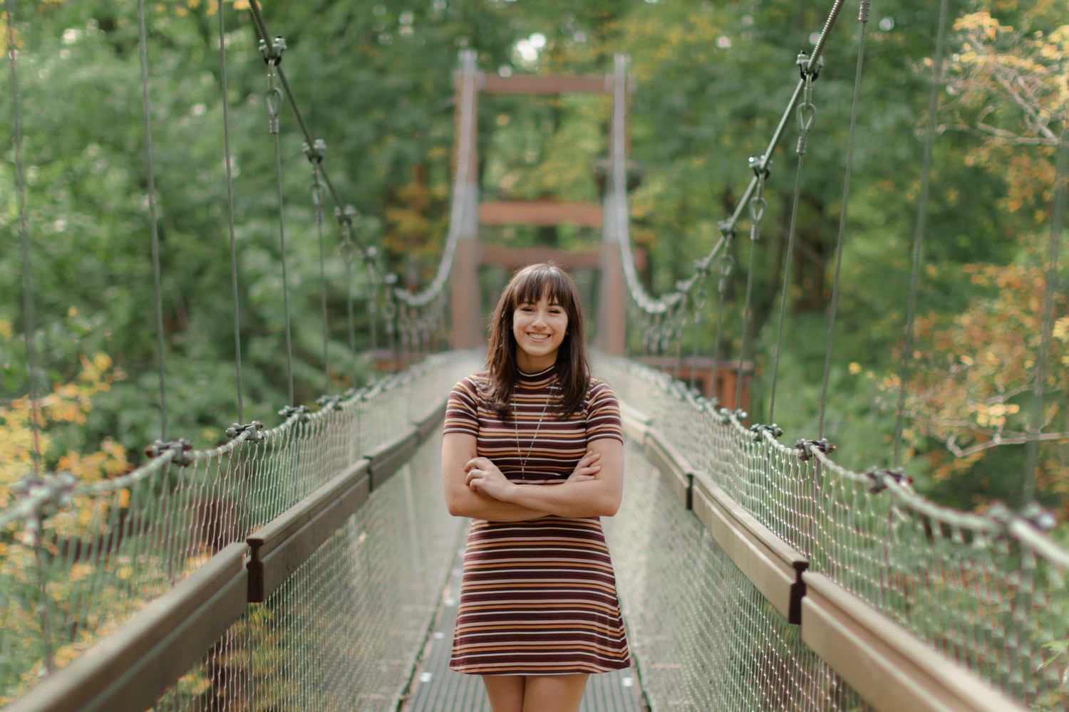 A person in a striped dress stands on a wooden suspension bridge surrounded by lush green forest.