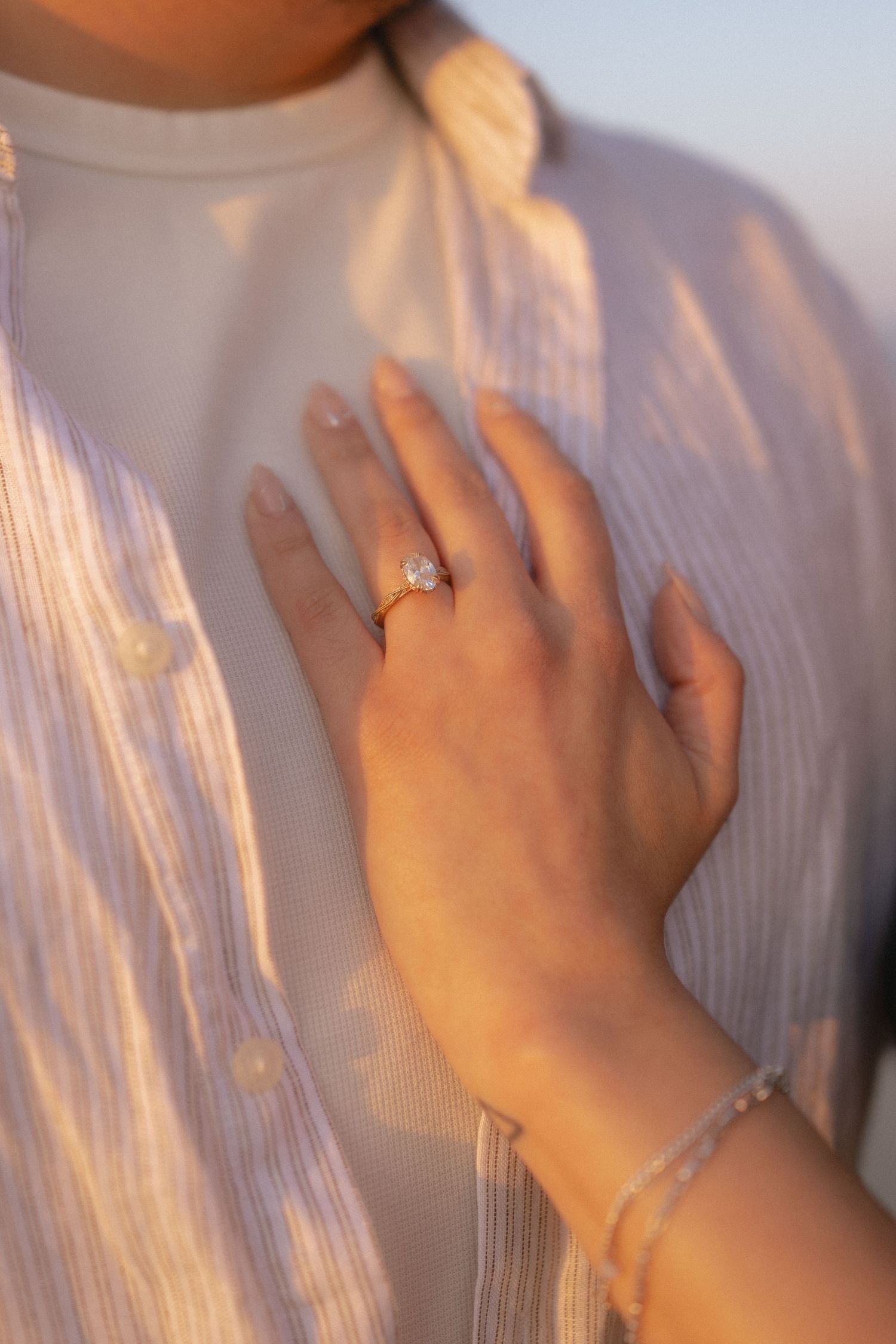 Close-up of a hand wearing a gold diamond engagement ring and bracelet resting on a chest in soft golden light.