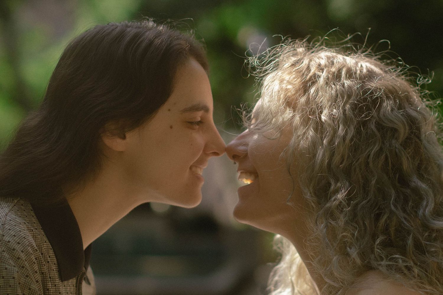 Two women sharing a tender moment, nearly kissing outdoors in soft natural light.