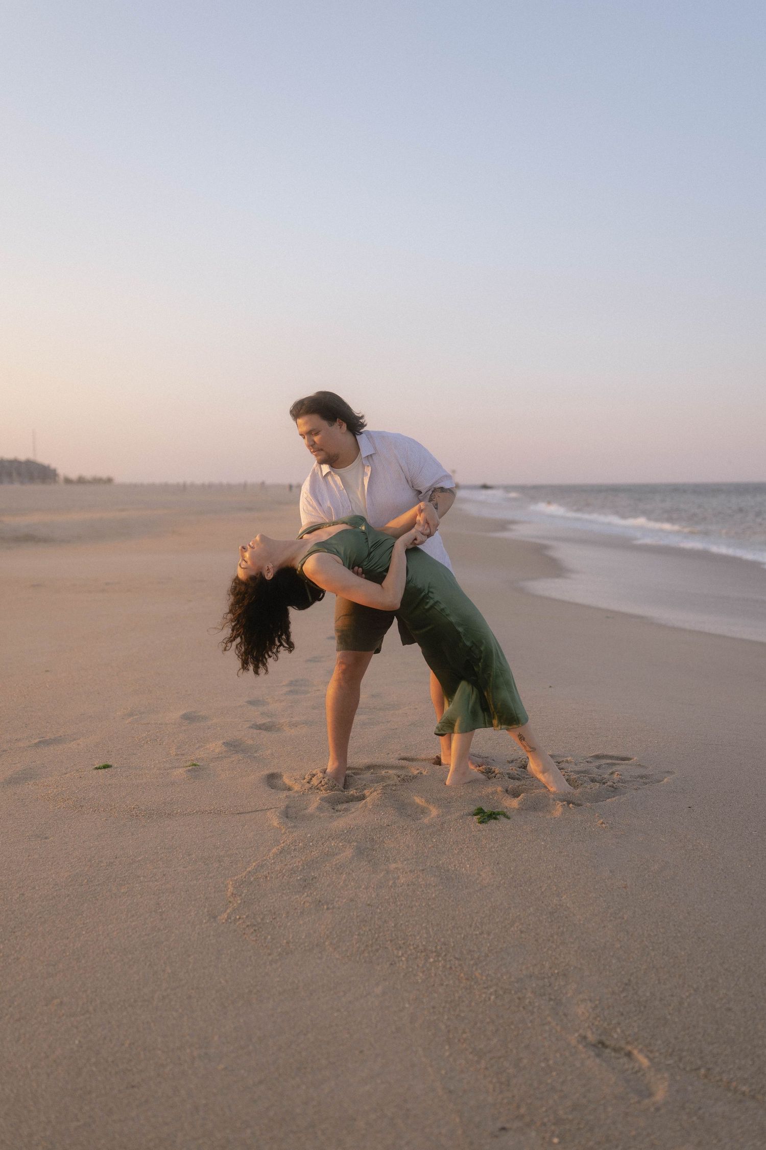 Couple dancing romantically on a sandy beach at sunset, woman in green dress dipping back gracefully.