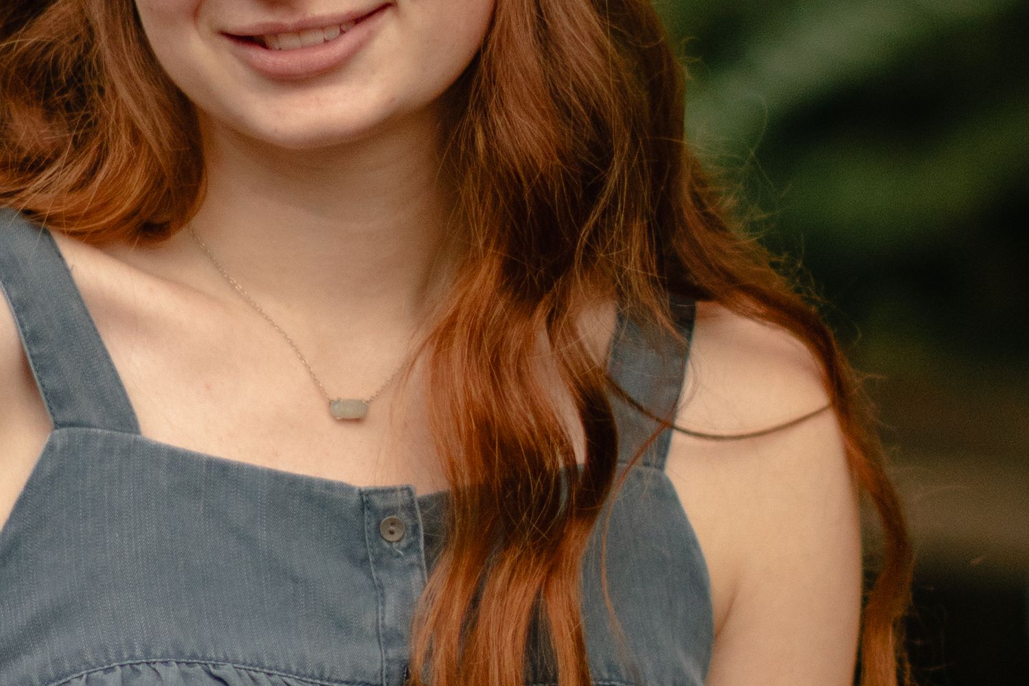 A close-up portrait shot of someone with auburn hair wearing a gray tank top against a blurred green background.