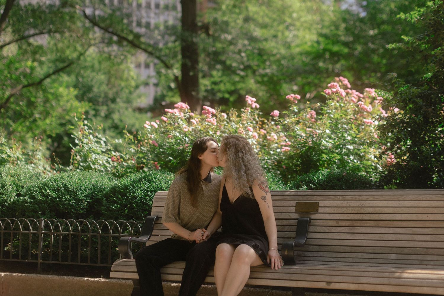 Two women sitting close together on a park bench surrounded by lush greenery and blooming pink roses.
