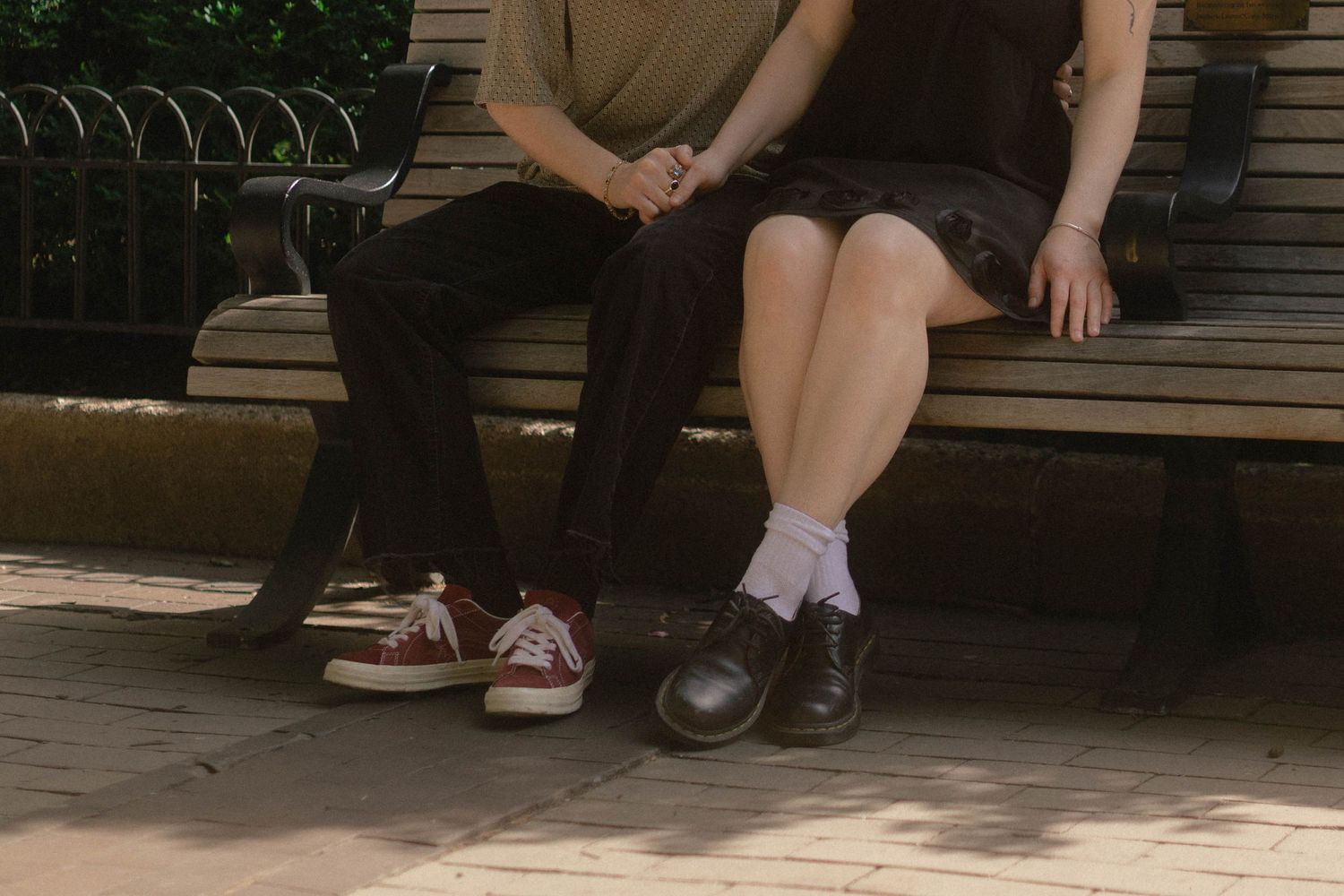 Two people sitting close together on a park bench, wearing casual shoes, one in red Converse and one in black loafers.