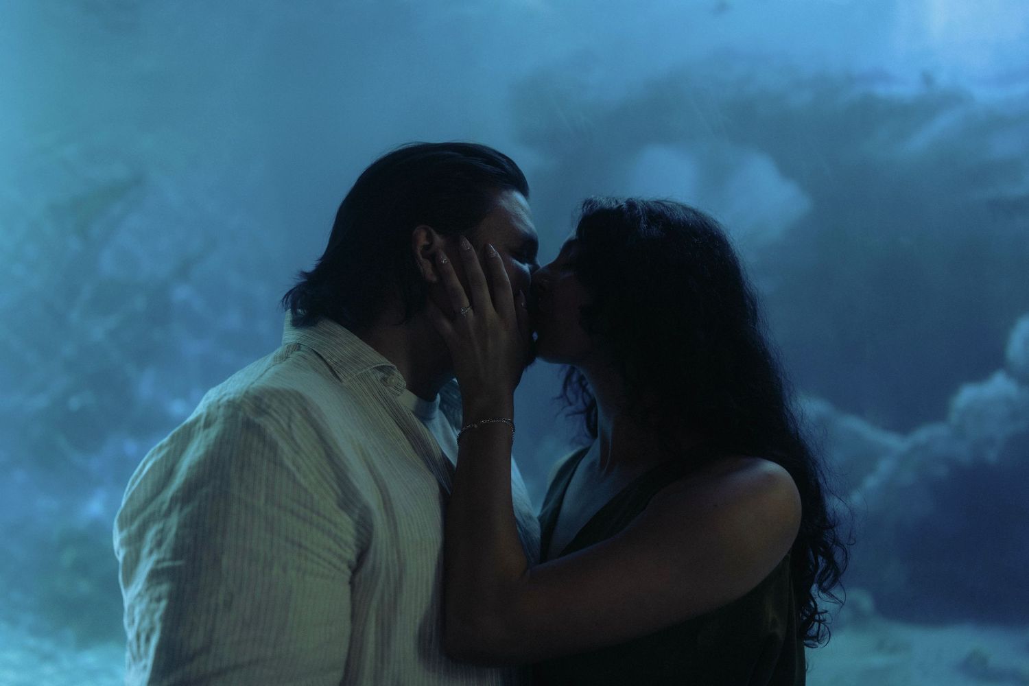 A romantic couple shares a passionate kiss in a dreamy blue-lit underwater-themed setting.