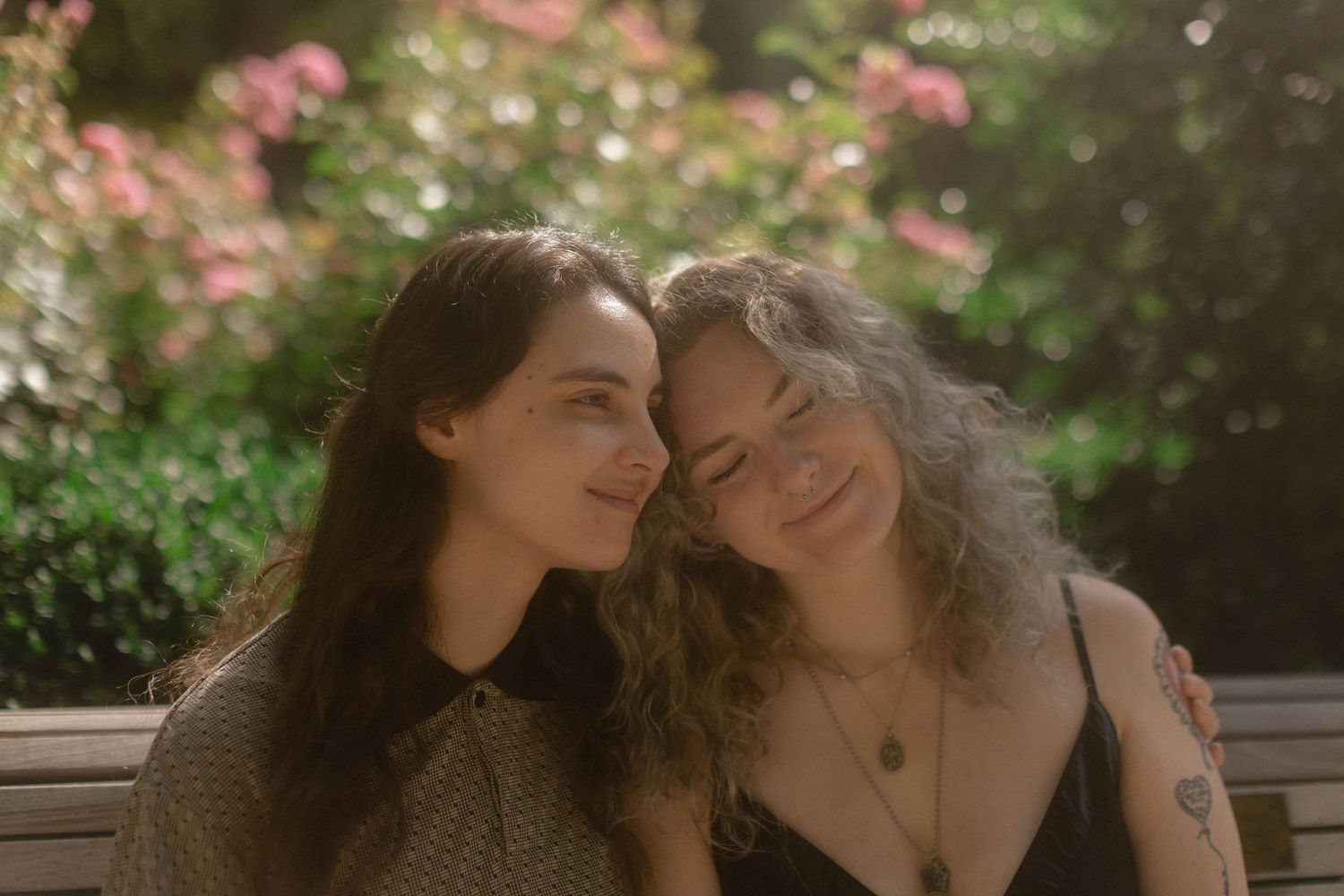Two women sitting close together on a bench in a sunlit garden with pink flowers in the background.