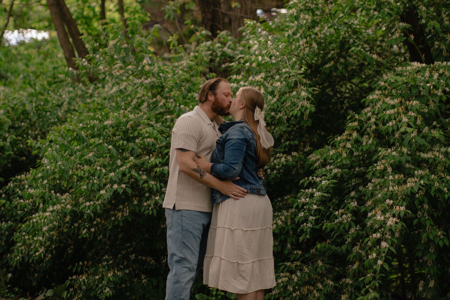 Couple sharing a romantic kiss outdoors surrounded by lush green flowering bushes in a garden setting.