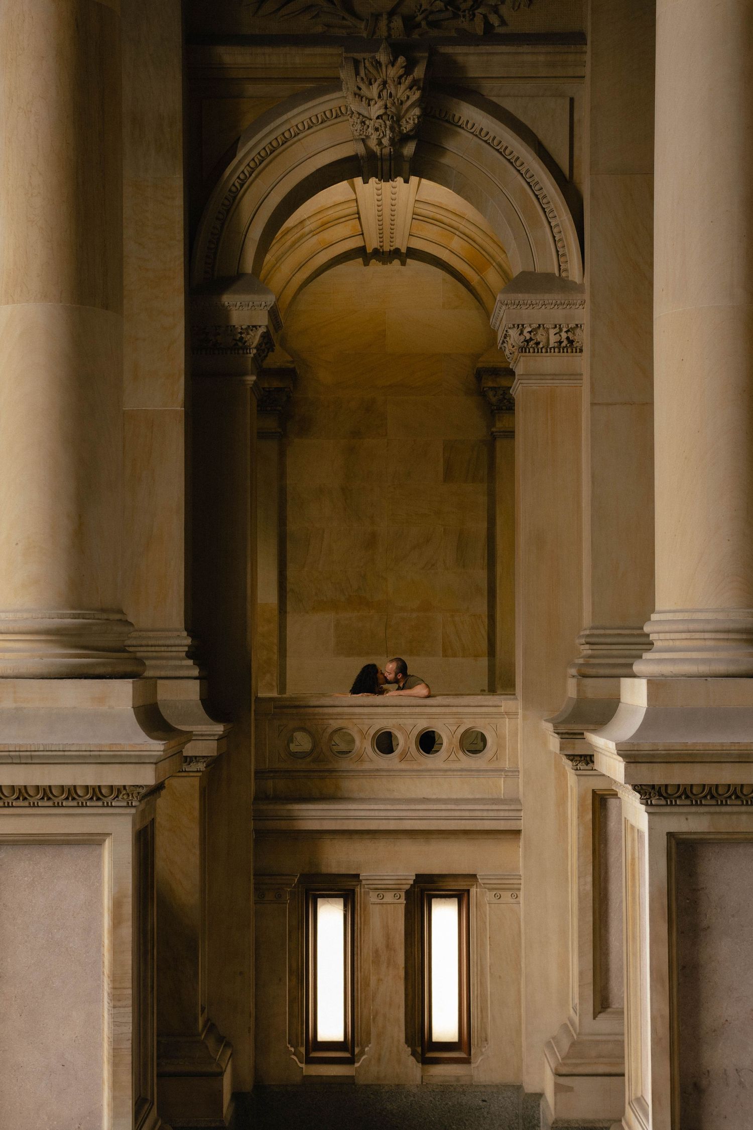 Person leaning over a balcony inside a grand neoclassical building with ornate arches and illuminated panels.
