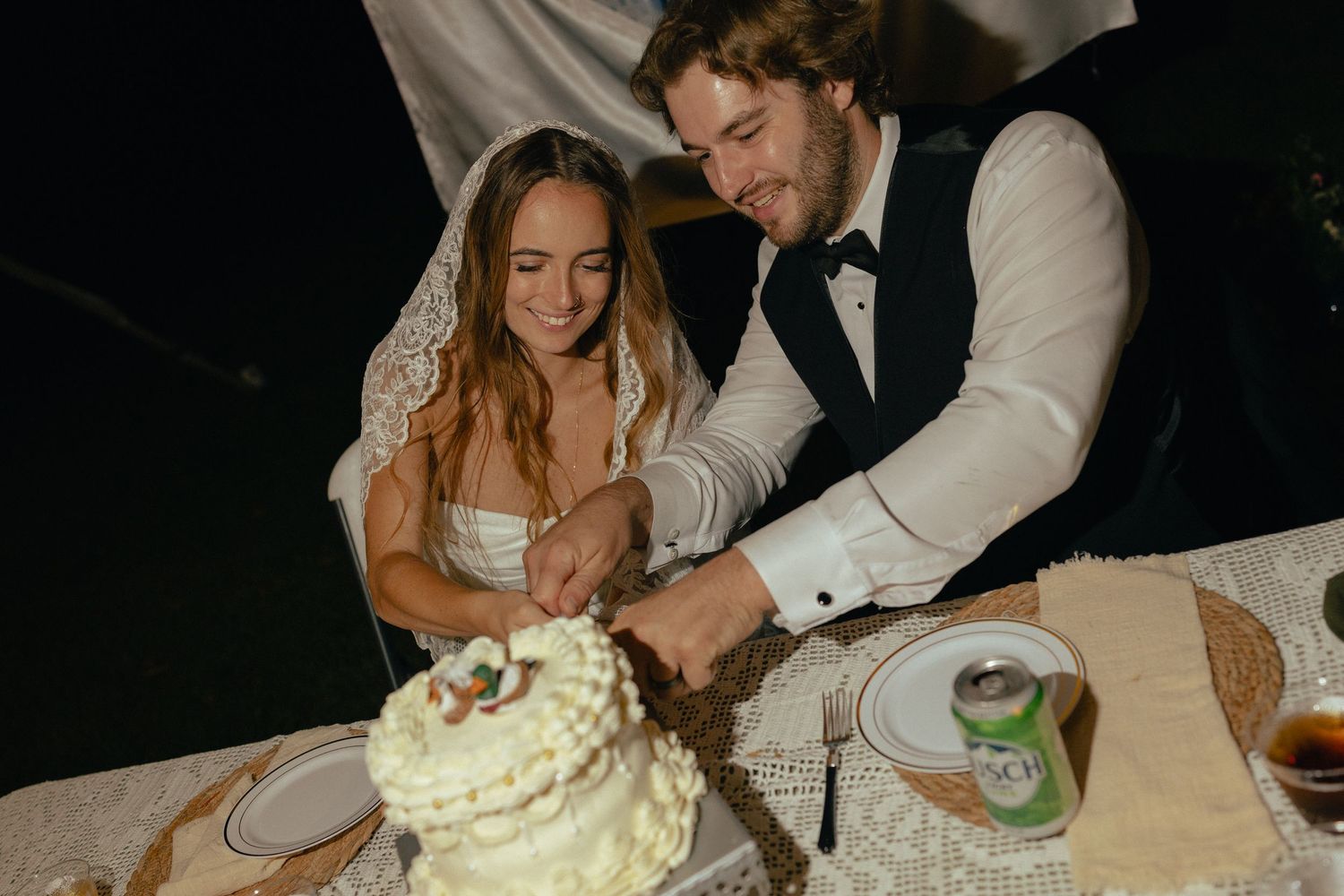 Bride and groom cutting white wedding cake together at outdoor evening reception table.
