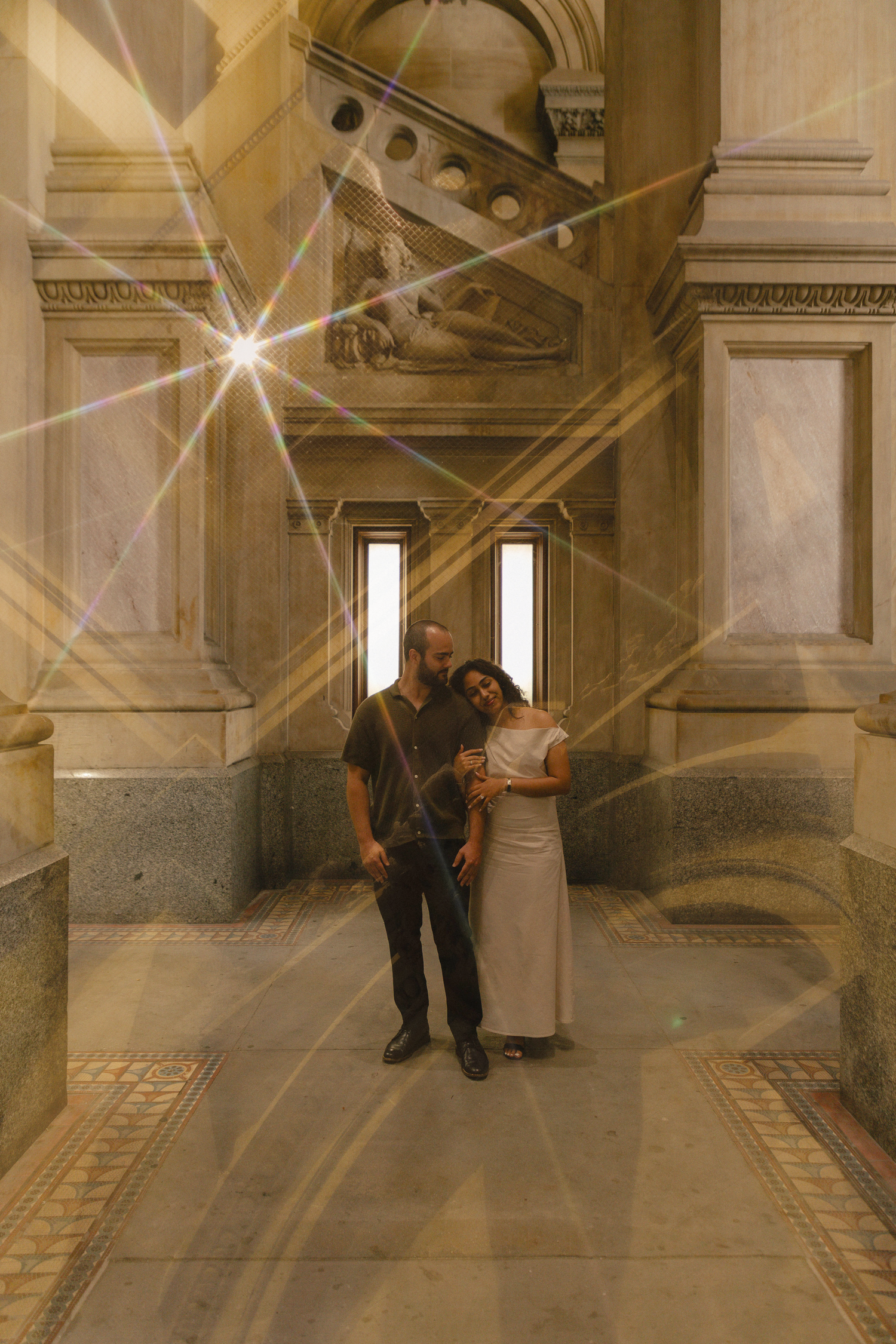 Couple embracing in an ornate marble hallway with elegant columns and a stunning lens flare effect.