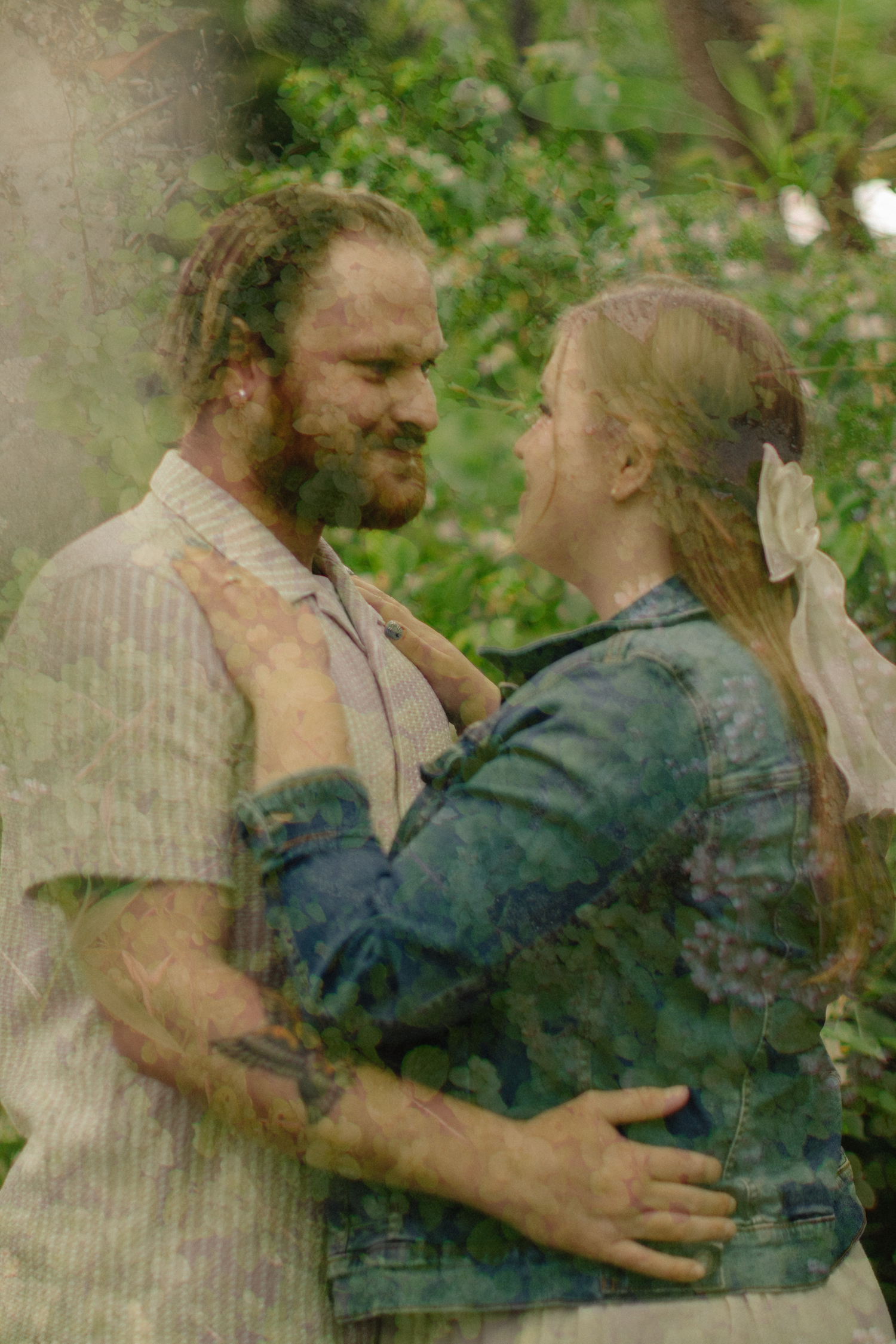 A bearded man with dreadlocks embraces a blonde woman in a denim jacket outdoors among lush green foliage.