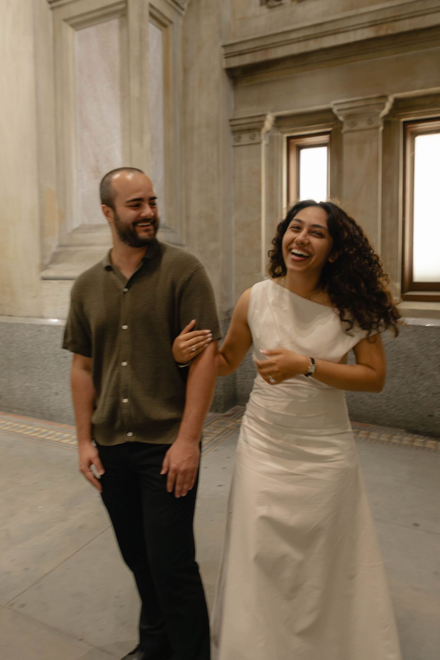 A laughing couple walks arm-in-arm inside an elegant building with marble columns, woman in white dress, man in olive shirt.