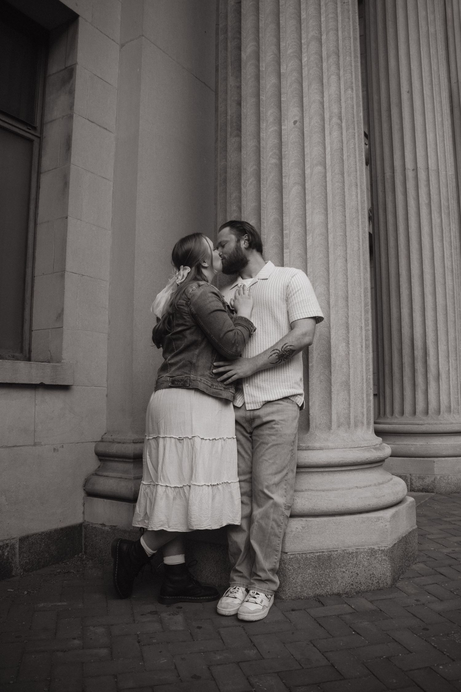 Black and white photo of a couple kissing beside stone columns of a historic building on a brick pathway.