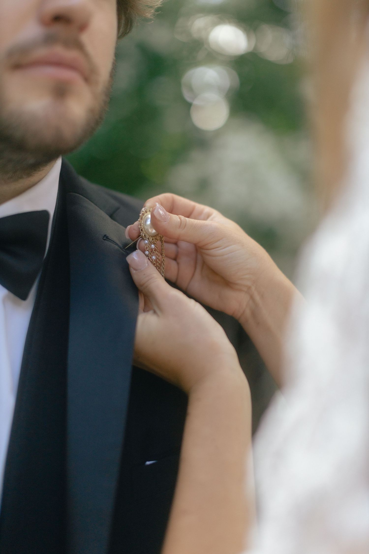 Bride pinning a gold boutonniere on groom's black tuxedo lapel during wedding preparation outdoors.