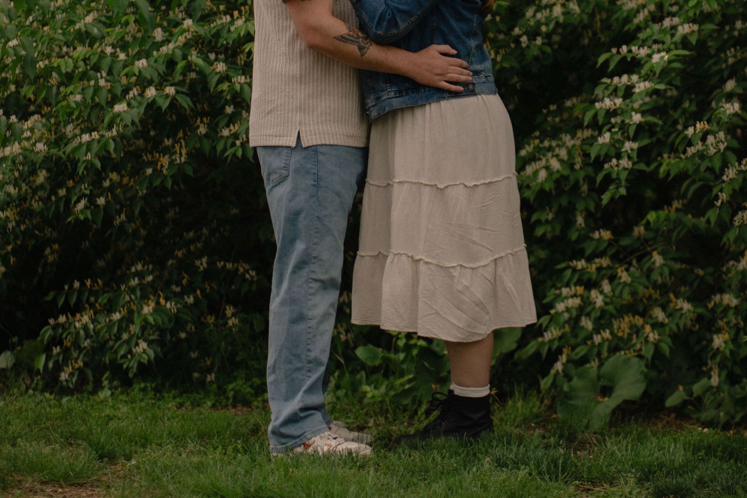 Couple embracing outdoors in a garden, wearing casual attire, standing on green grass near lush bushes.
