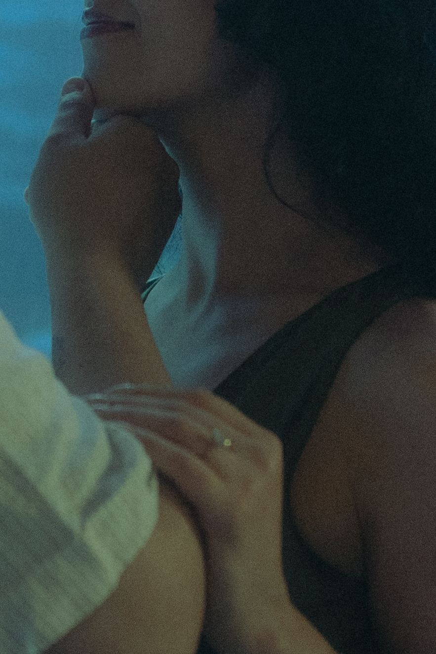 A woman in a dark top poses thoughtfully in moody blue lighting while someone rests a hand on her shoulder.