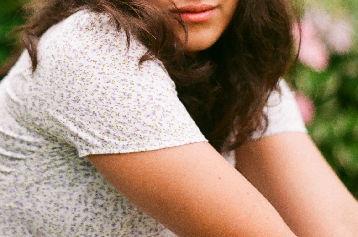 Young woman with curly dark hair wearing a floral print top, sitting outdoors near green foliage.