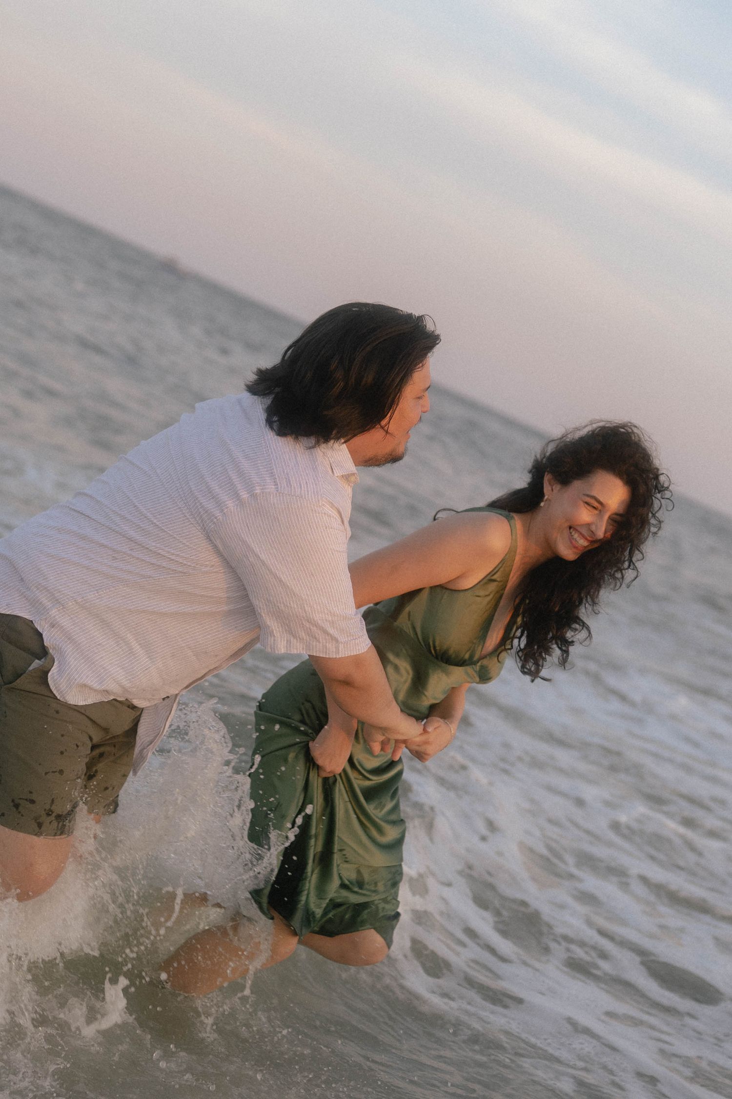 Couple laughing and splashing in ocean waves at sunset, woman in green dress held by man in white shirt.