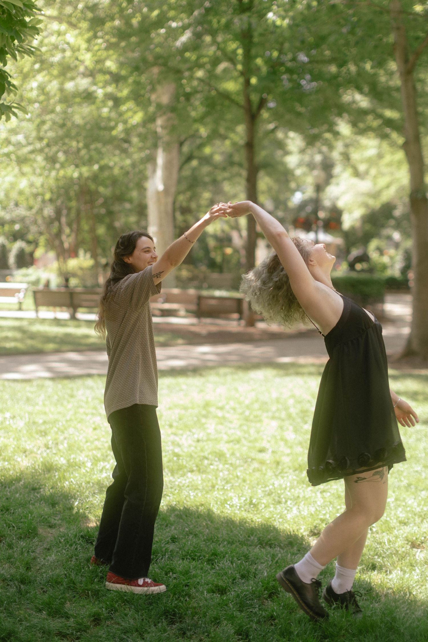 Two women dancing together on green grass in a sunny park, holding hands raised above their heads.