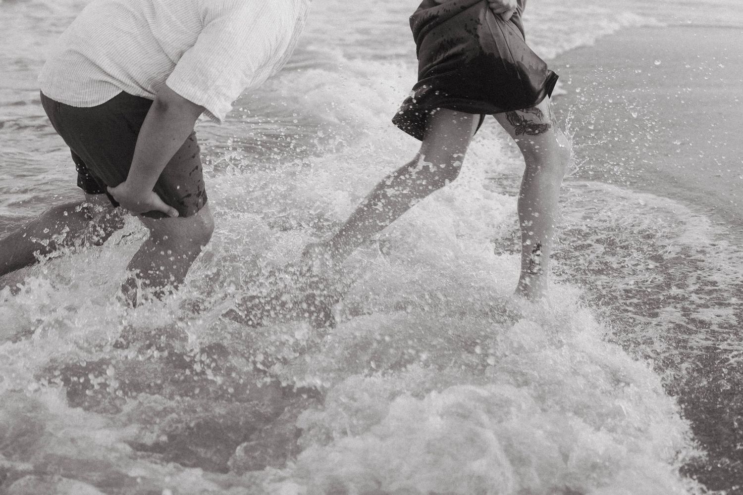 Two people splashing through ocean waves at the beach in a black and white photo.