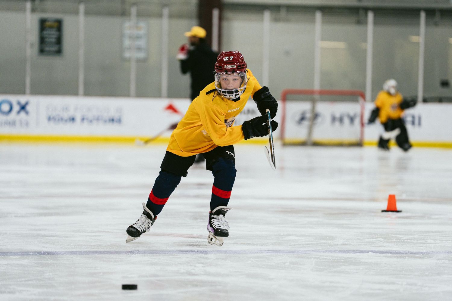 Young hockey player in yellow jersey practicing puck control around orange cone during ice skating drill.