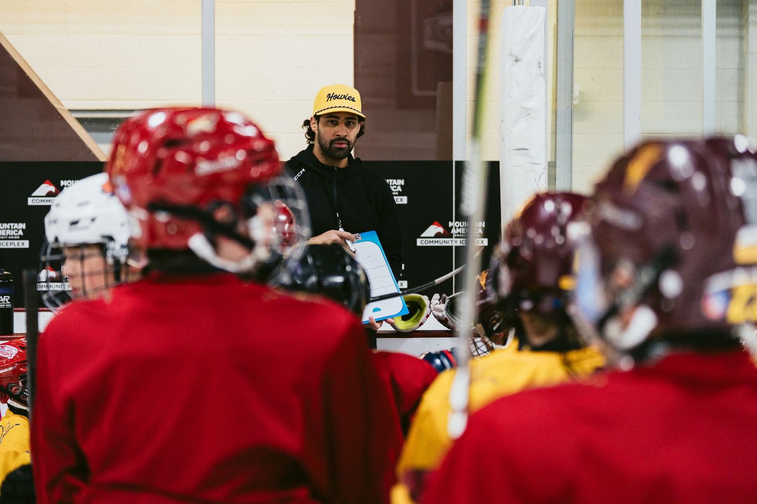 Hockey coach in black jacket addresses team of young players wearing red and yellow jerseys during ice rink huddle.