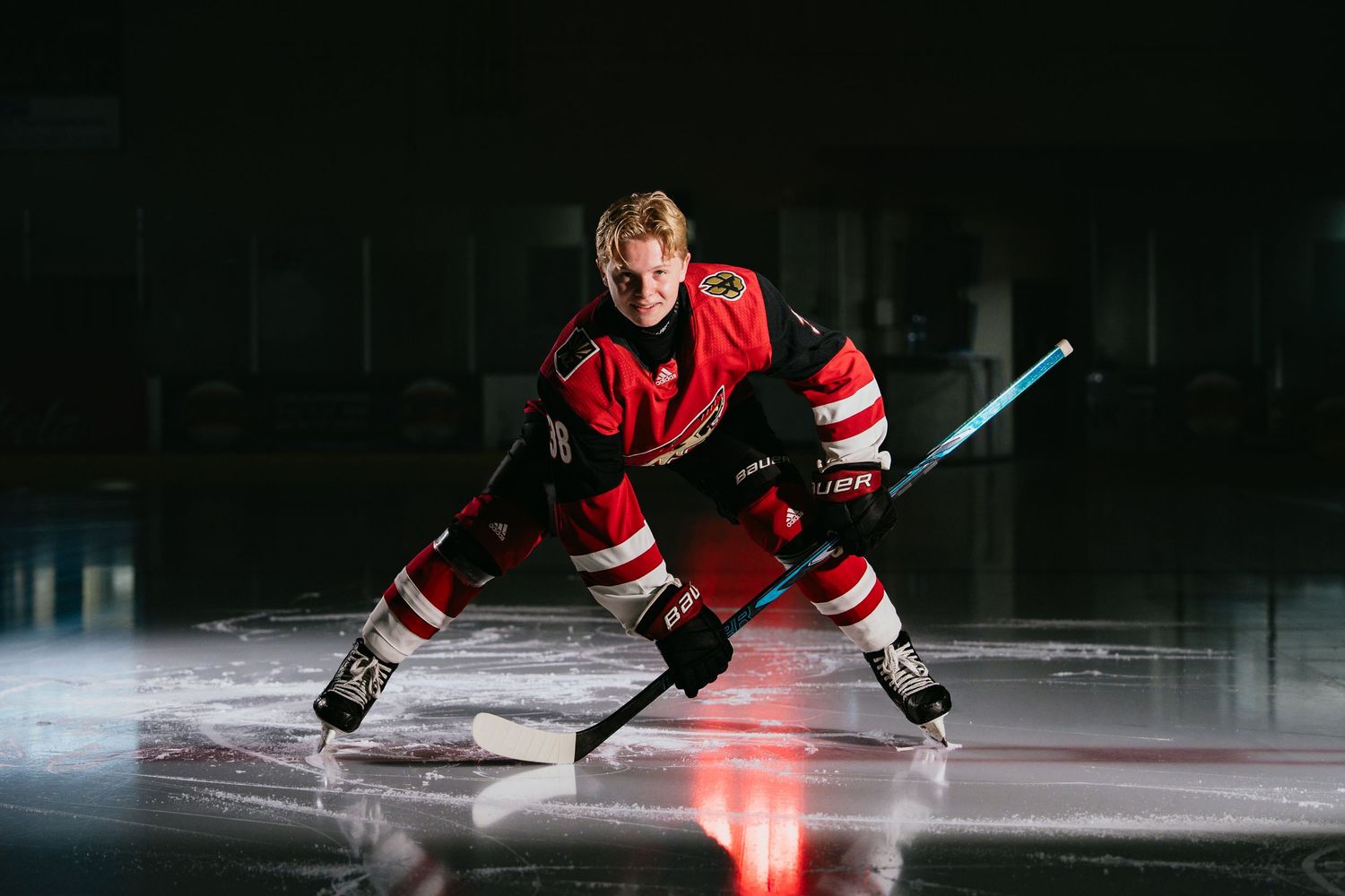 Hockey player in red jersey crouches on ice rink, holding stick in dramatic dark arena lighting.