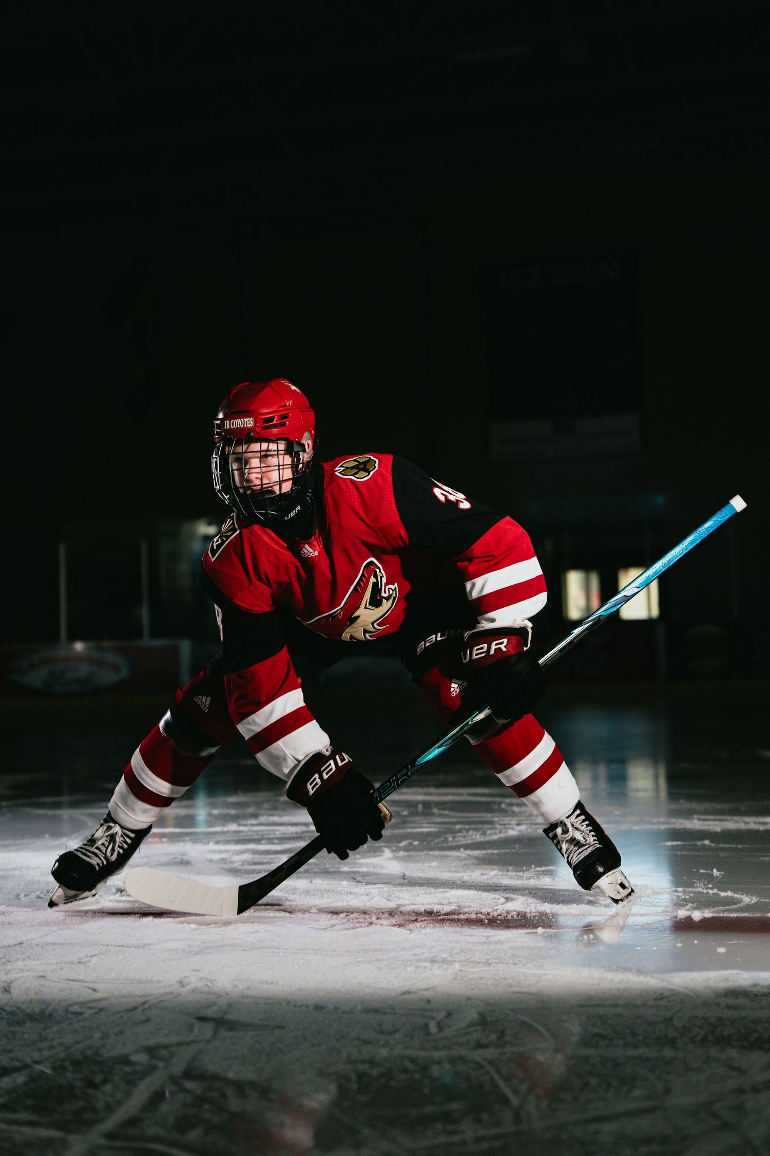 Young ice hockey player in red Arizona Coyotes jersey in ready position on dark rink.