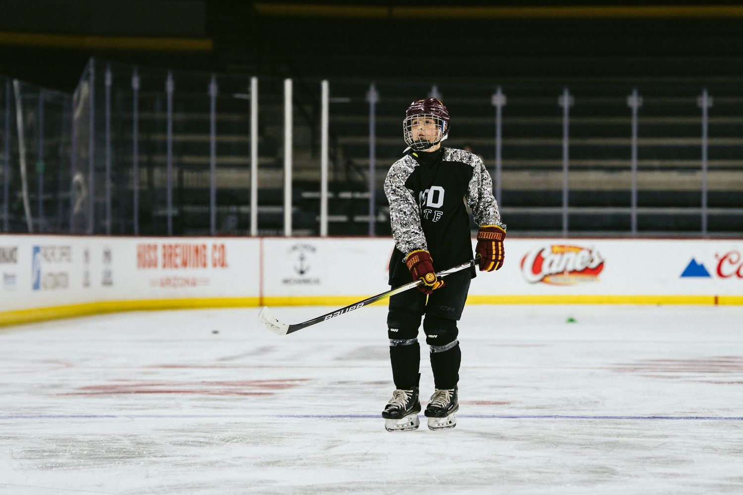 Young hockey player in camo uniform stands on the ice holding a stick during indoor practice.