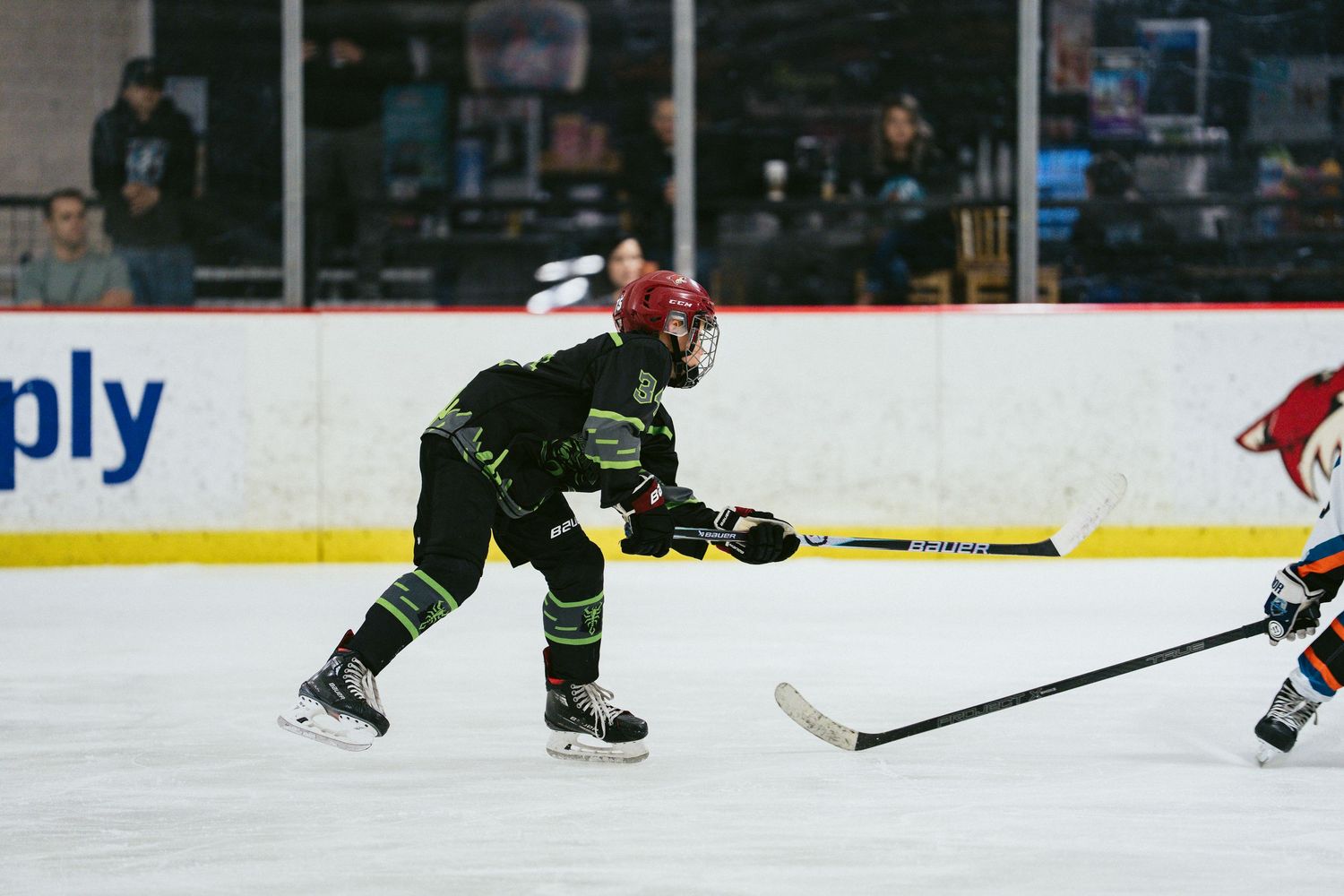 Youth hockey player in black and green uniform takes a shot on ice during an indoor hockey game.