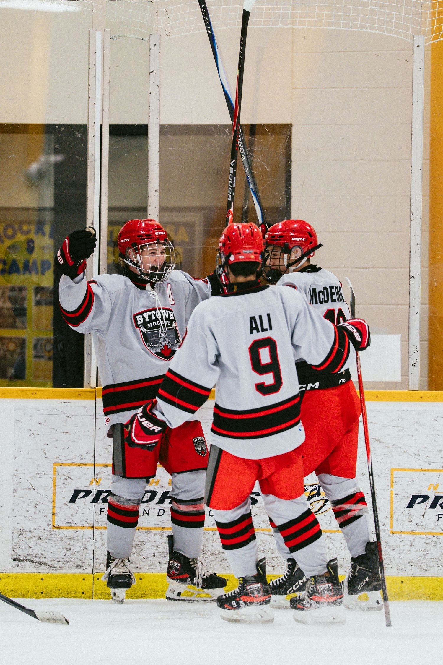 Youth hockey players in red and white jerseys celebrate a goal on the ice, raising sticks in triumph.