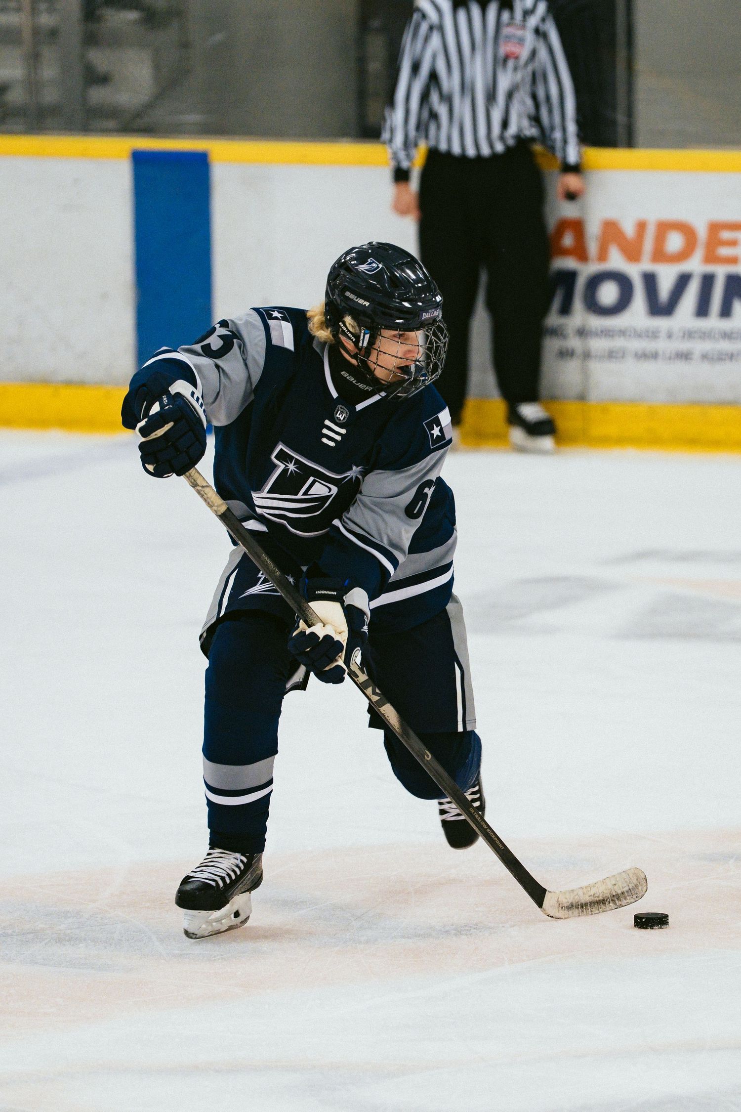 A youth hockey player in a navy uniform handles the puck on the ice with a referee in the background.