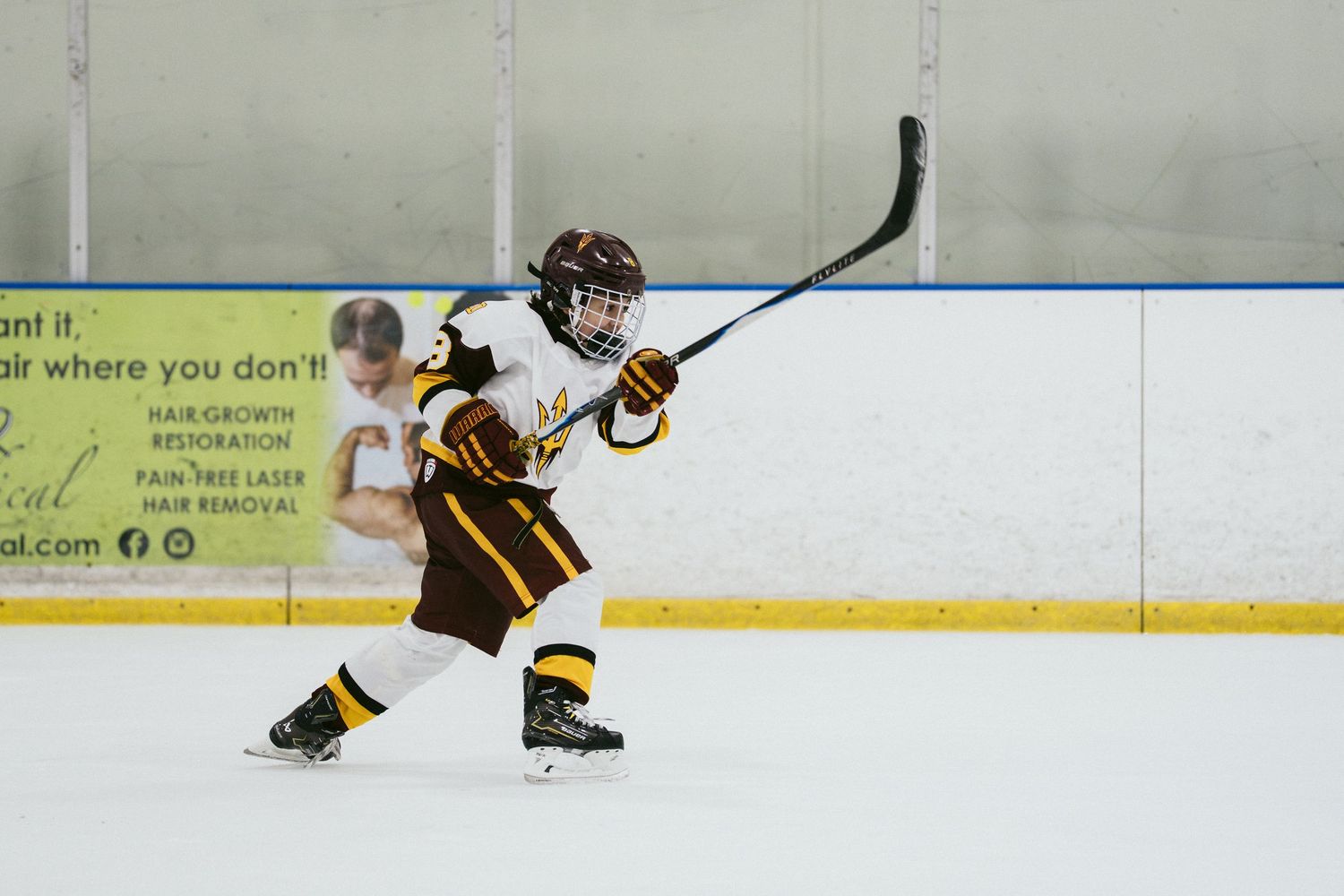 Young youth hockey player in maroon uniform taking a shot on ice during a hockey game at indoor rink.
