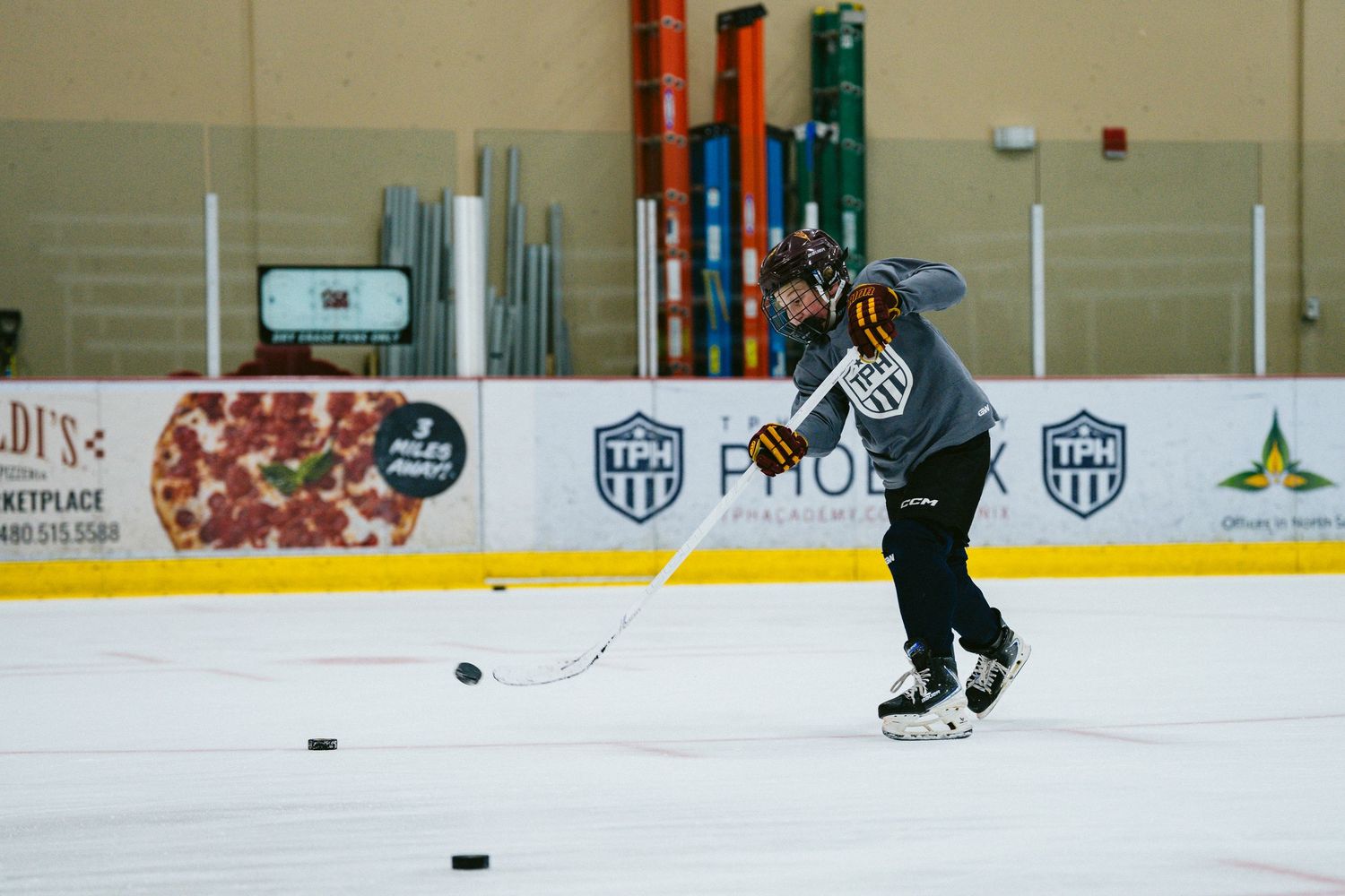 A hockey player shoots a puck on the ice during practice at an indoor rink with ads on the boards.
