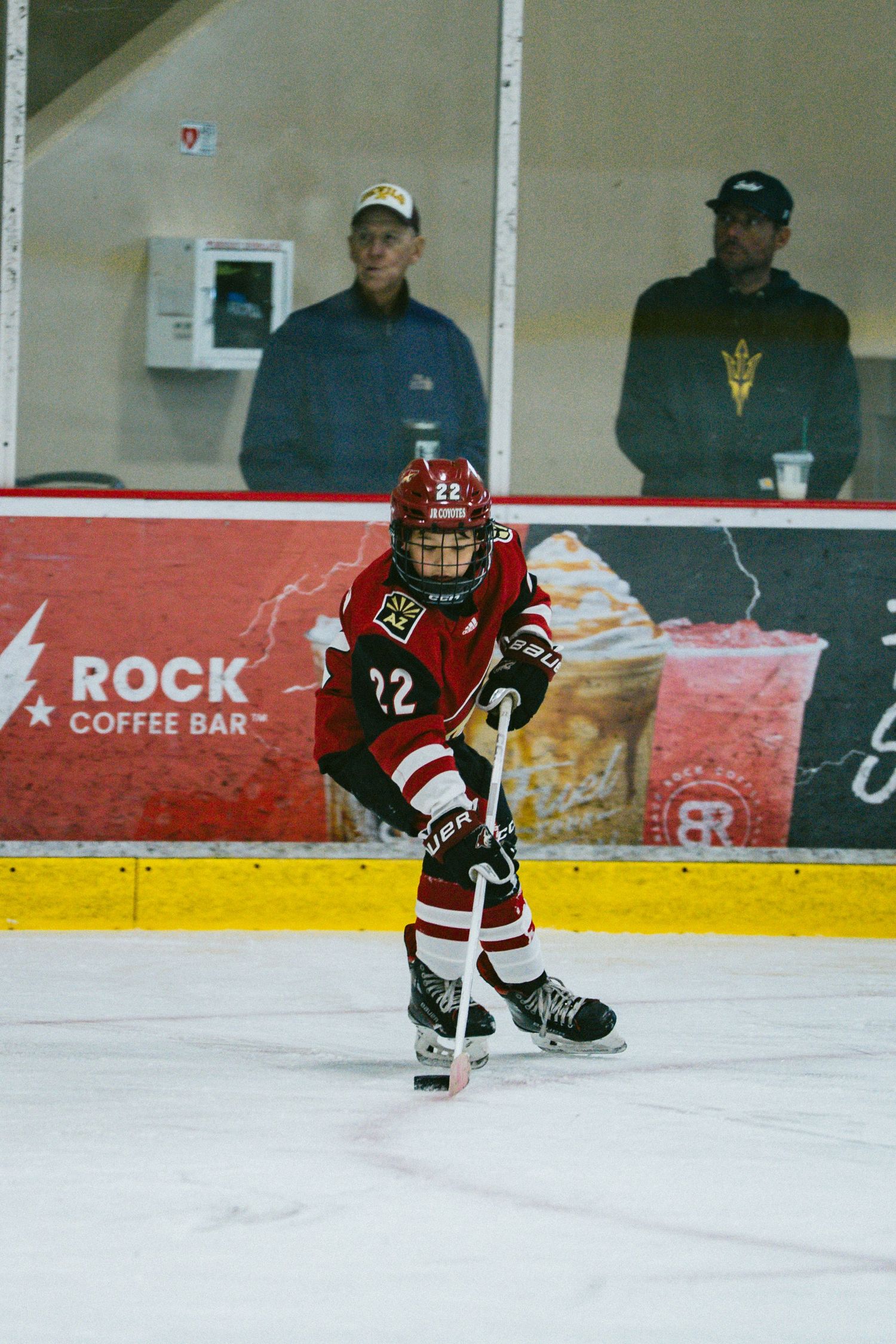 Youth hockey players in red and black jerseys skating on ice during a game near Rock Coffee Bar.