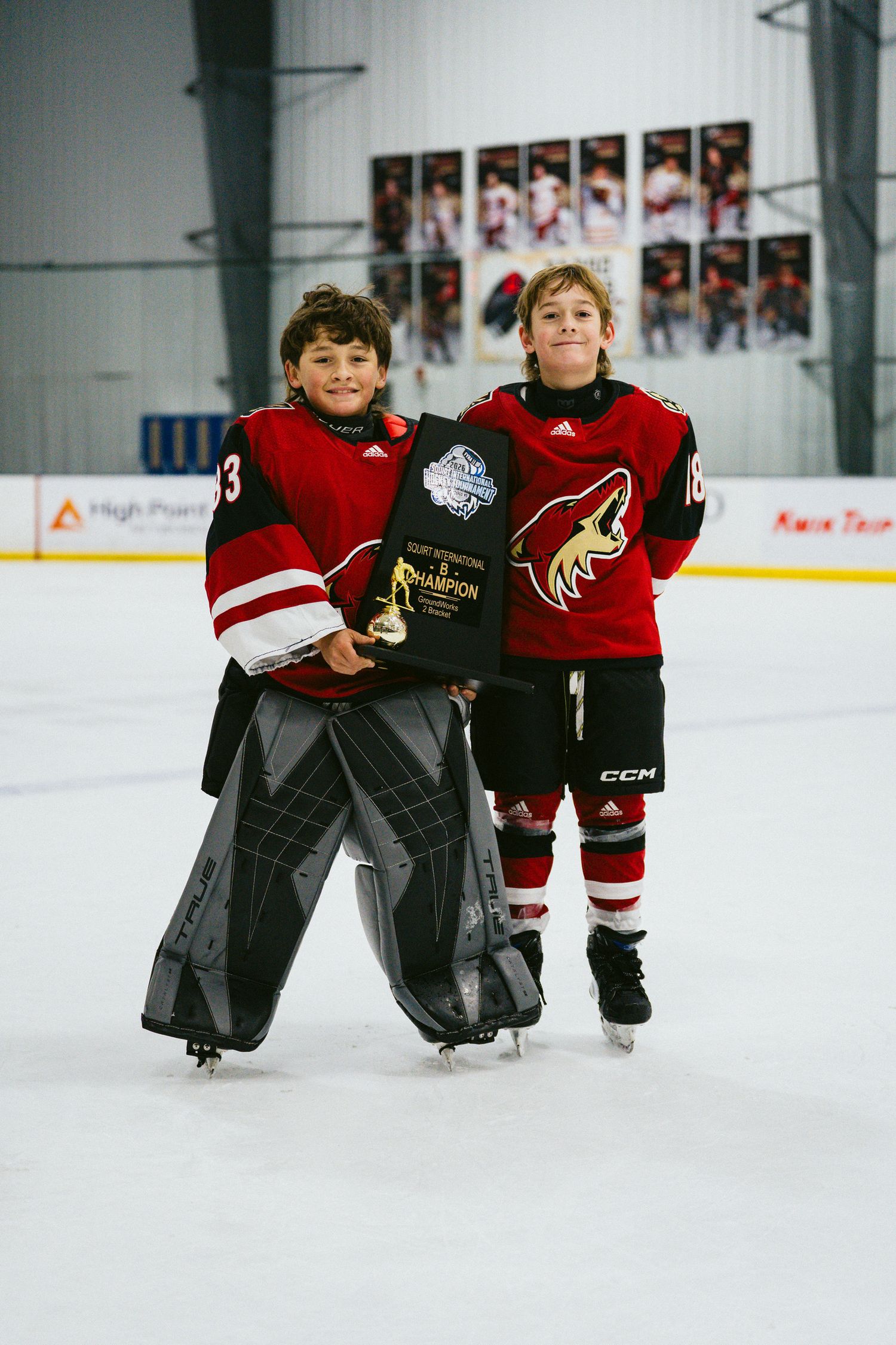 Hockey players in red jerseys pose with championship awards on indoor rink.