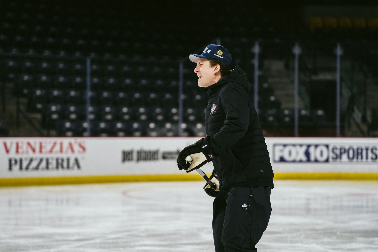A hockey coach skates on an indoor ice rink during practice at an arena with FOX 10 Sports signage.