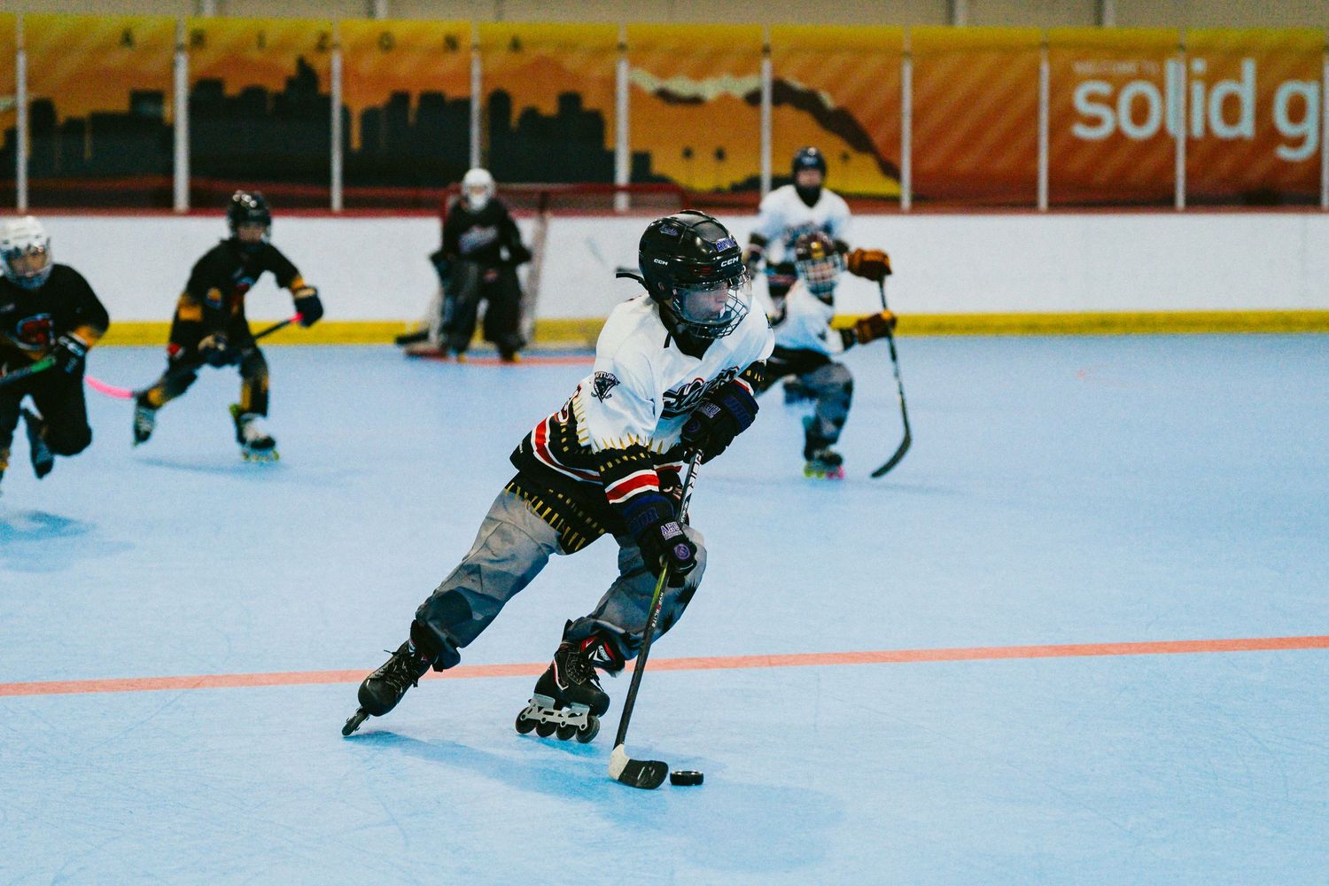 Players compete in an indoor inline roller hockey game on a smooth rink, with one player in focus controlling the puck.