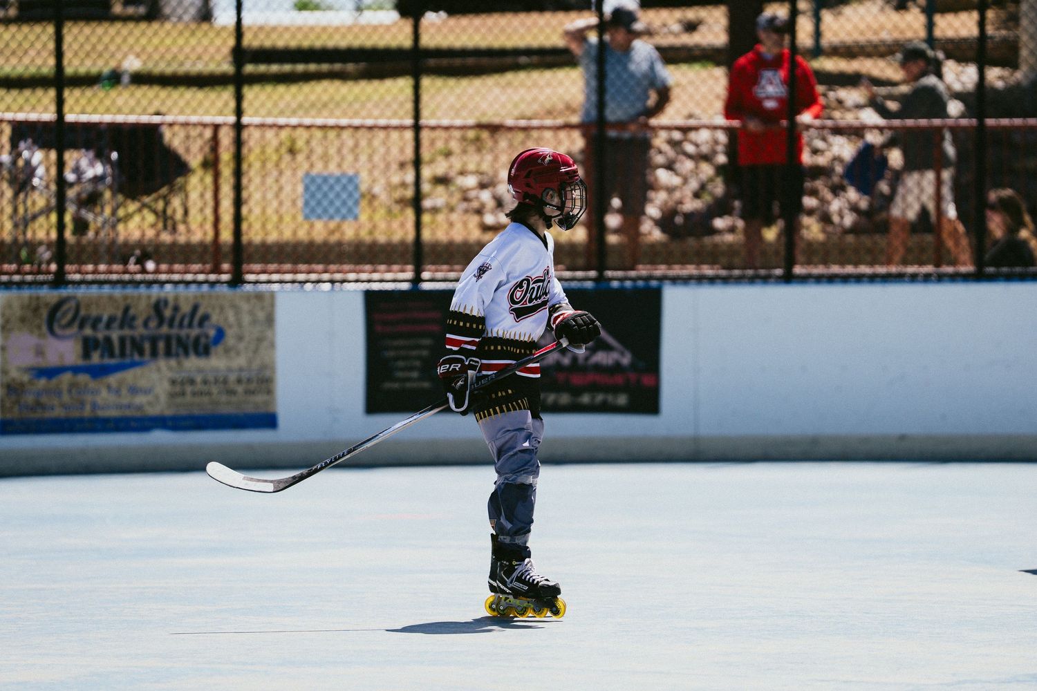 Youth roller hockey player in white Outlaws jersey skates with stick on outdoor rink during sunny game.