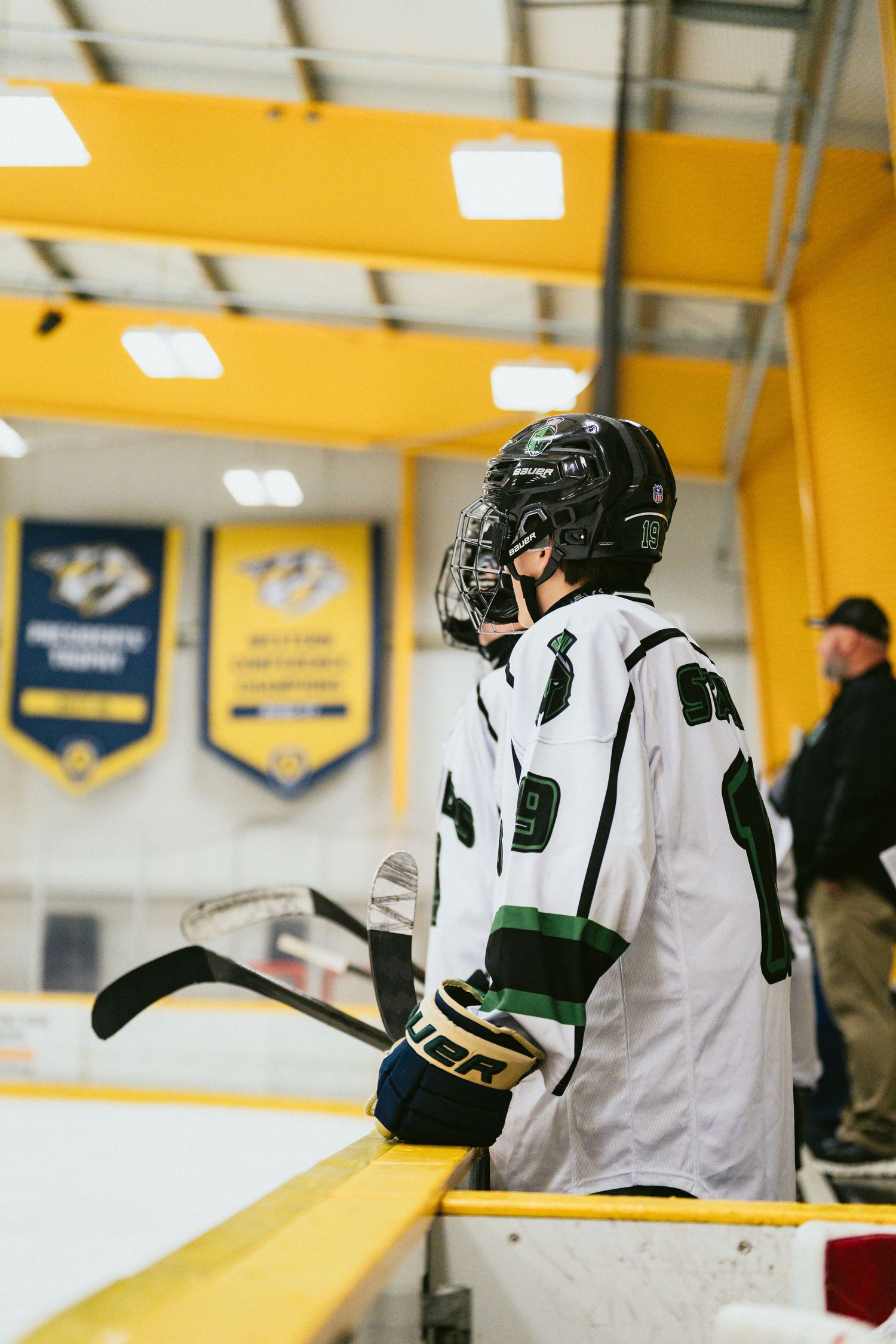 Young hockey player in white and green jersey watches game from the bench at an indoor ice rink.