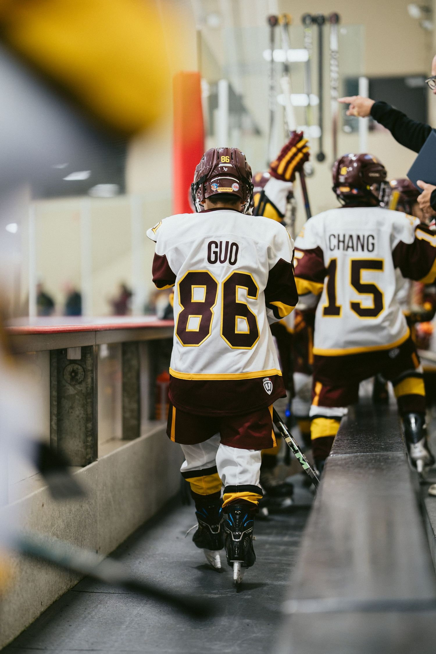 Youth hockey players in white and maroon jerseys walk through a tunnel toward the ice rink.