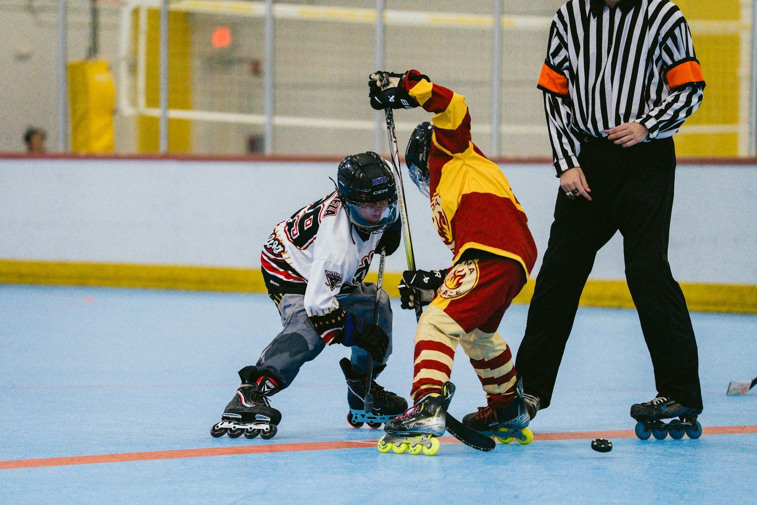 Two youth roller hockey players face off for a puck drop as a referee watches during an indoor rink game.