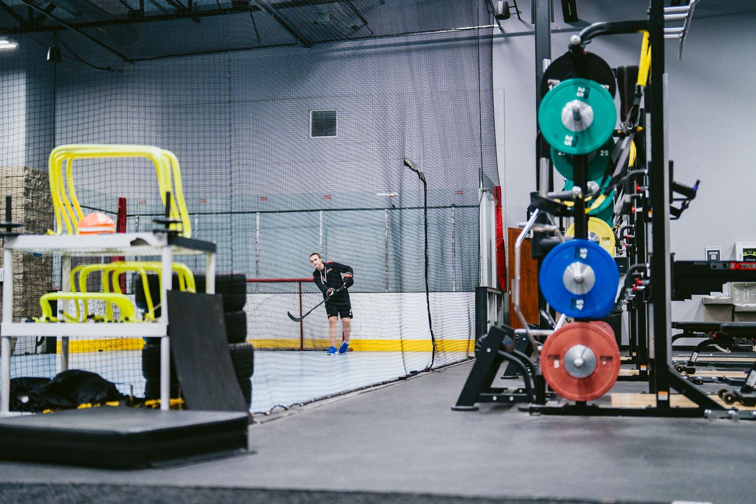 Hockey training facility with weight equipment, colorful bumper plates, and a player on the ice rink in the background.