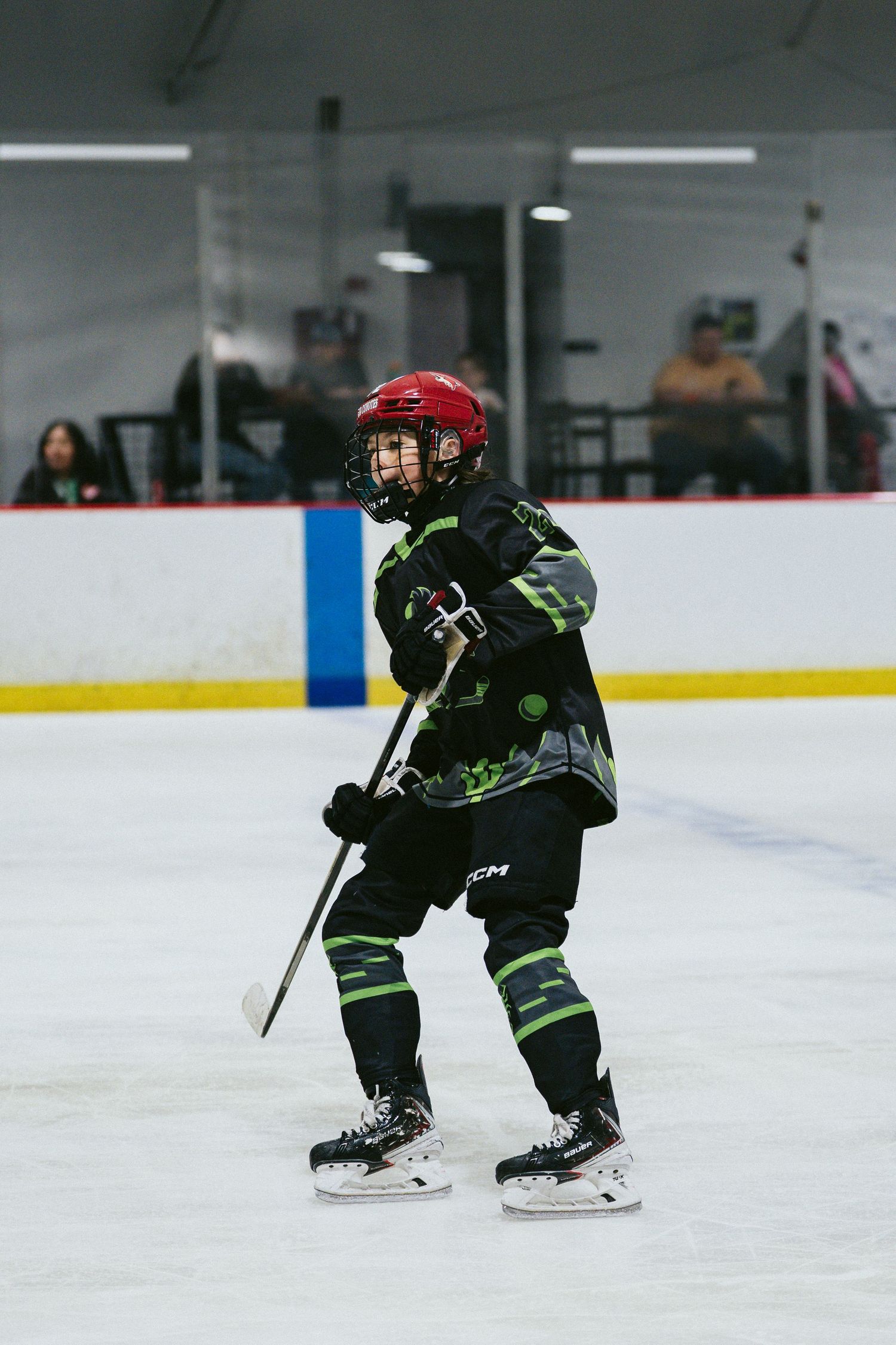 Young hockey player in black green uniform and red helmet stands ready on indoor ice rink.