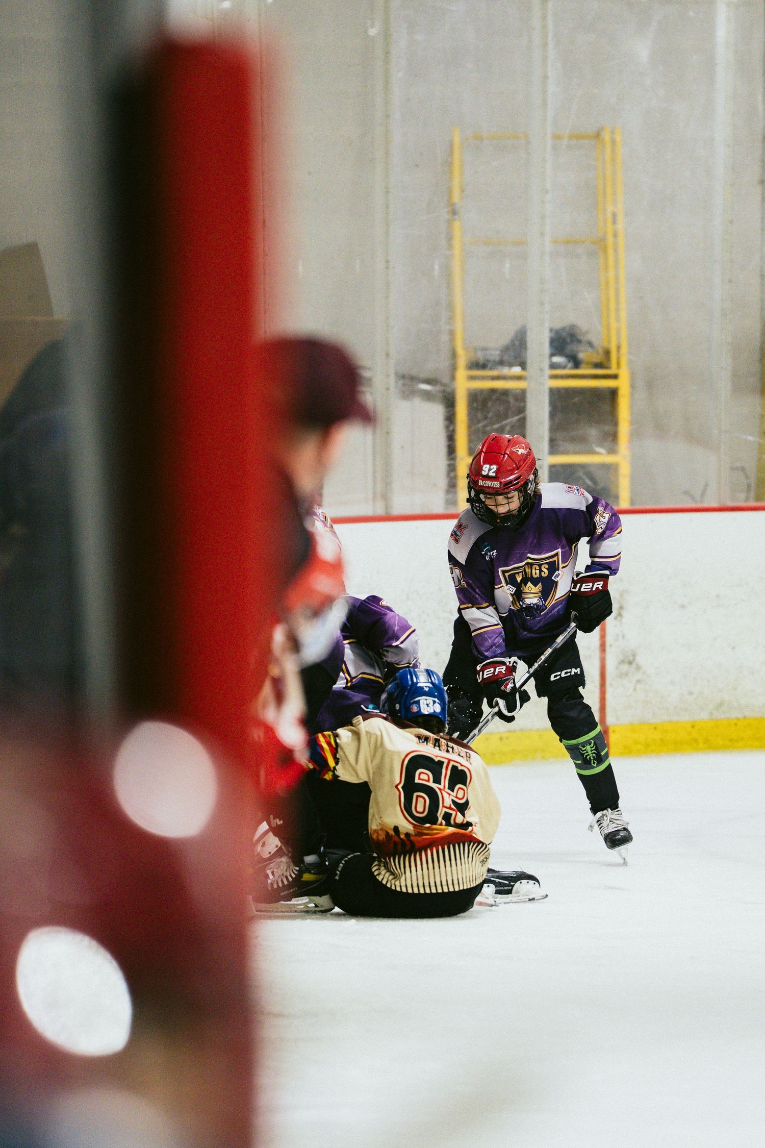 Youth hockey player in purple uniform skates near a fallen opponent during an indoor ice hockey game.